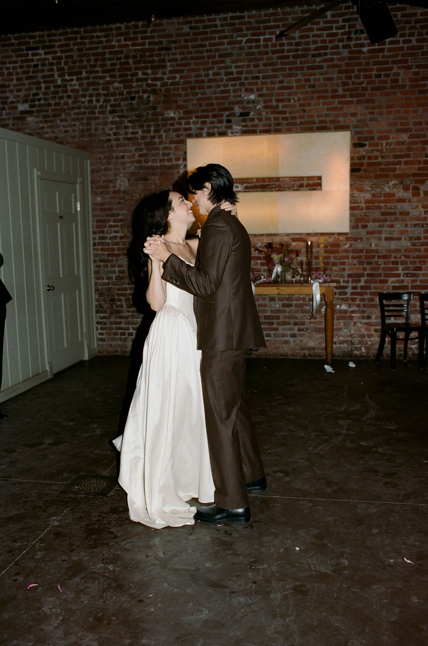 A couple in formal wedding attire share an intimate dance moment against an exposed brick wall background.