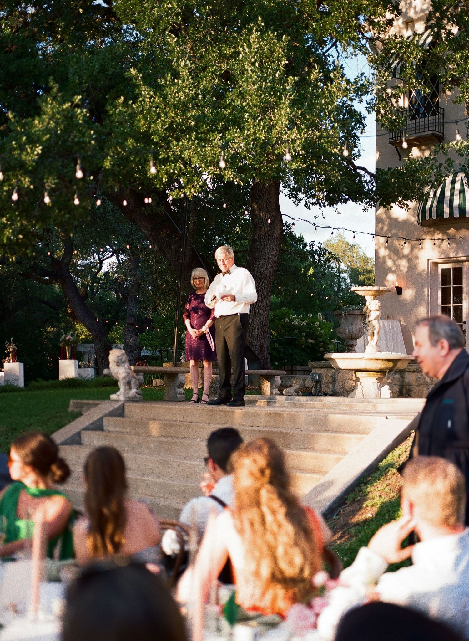 Outdoor wedding reception guests seated at tables under trees and string lights.