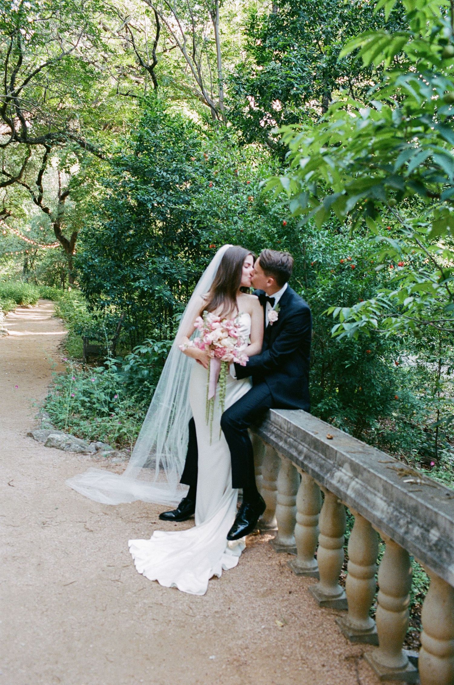A romantic moment captured on a garden path with lush greenery and stone railings.