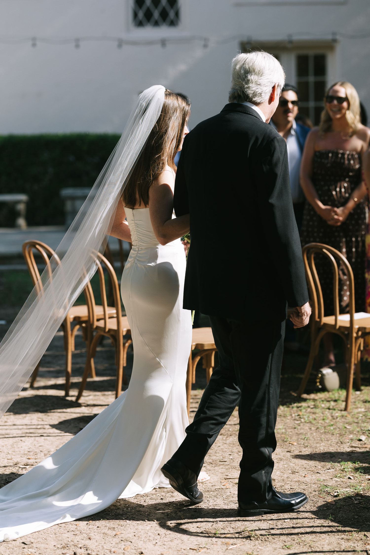 Flowing white wedding dress and veil trailing on garden path during outdoor ceremony.
