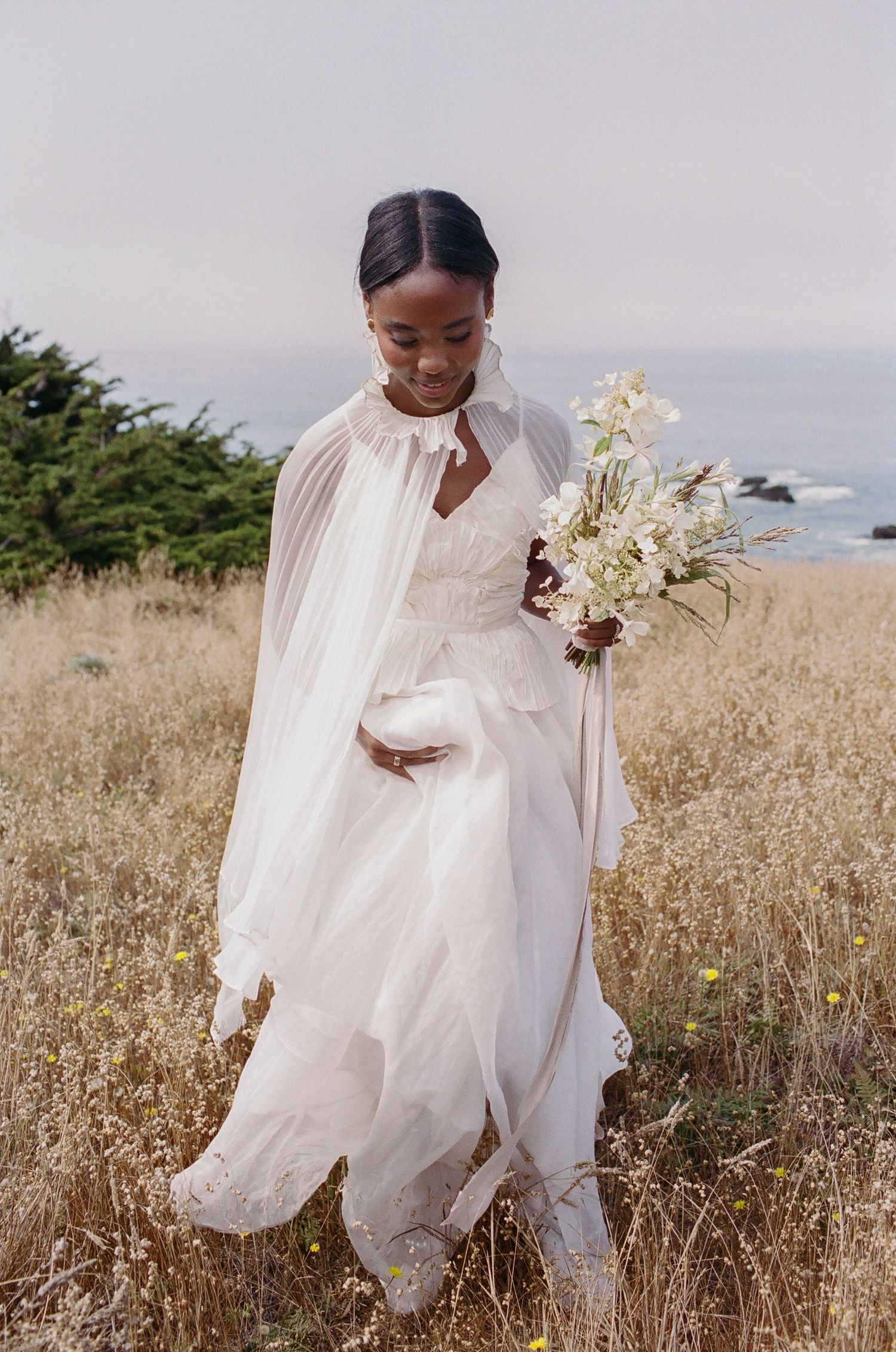 A flowing white wedding dress billows in the wind against a scenic coastal meadow backdrop.