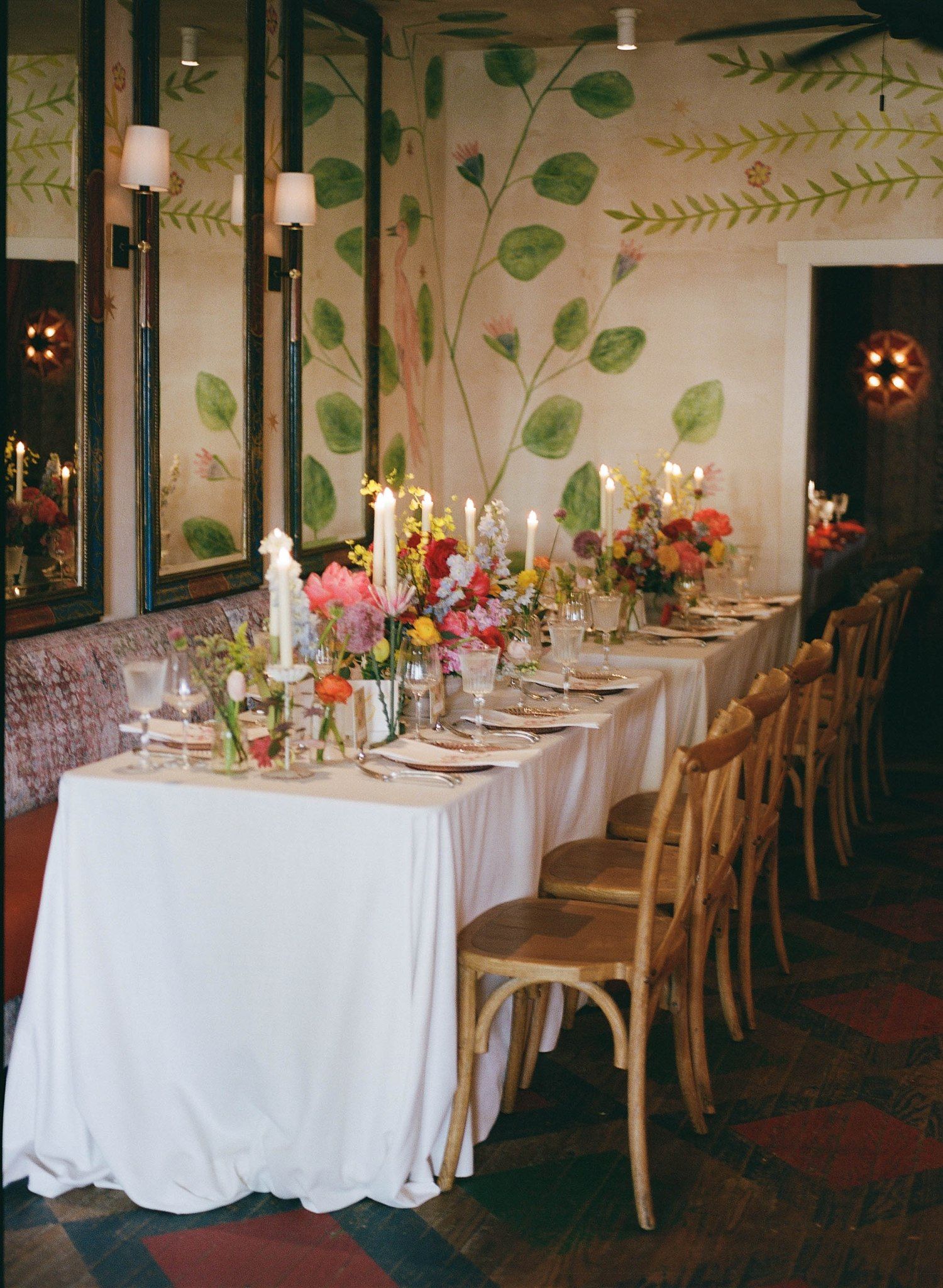 Wedding reception tables with white linens and wooden chairs arranged in room with painted wall mural.