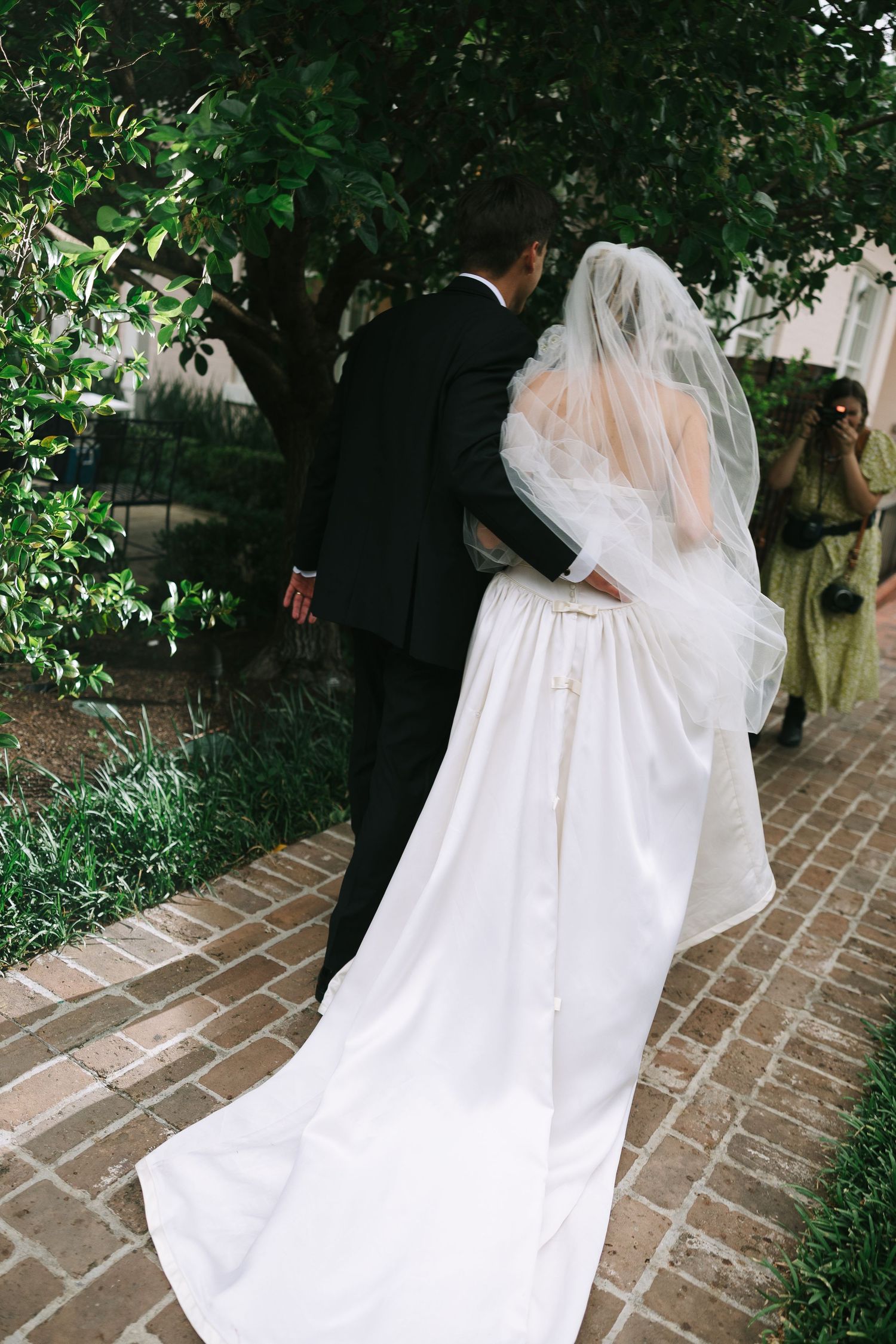 White wedding dress with long train and veil trailing along a brick pathway surrounded by greenery.