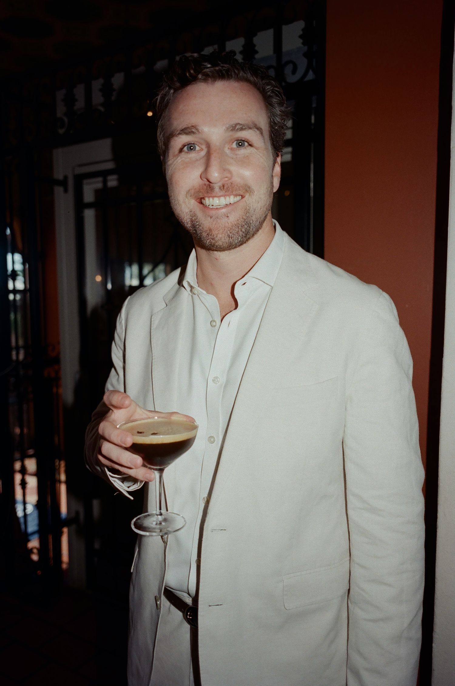 Person in a white suit jacket holds a wine glass in a dark room.