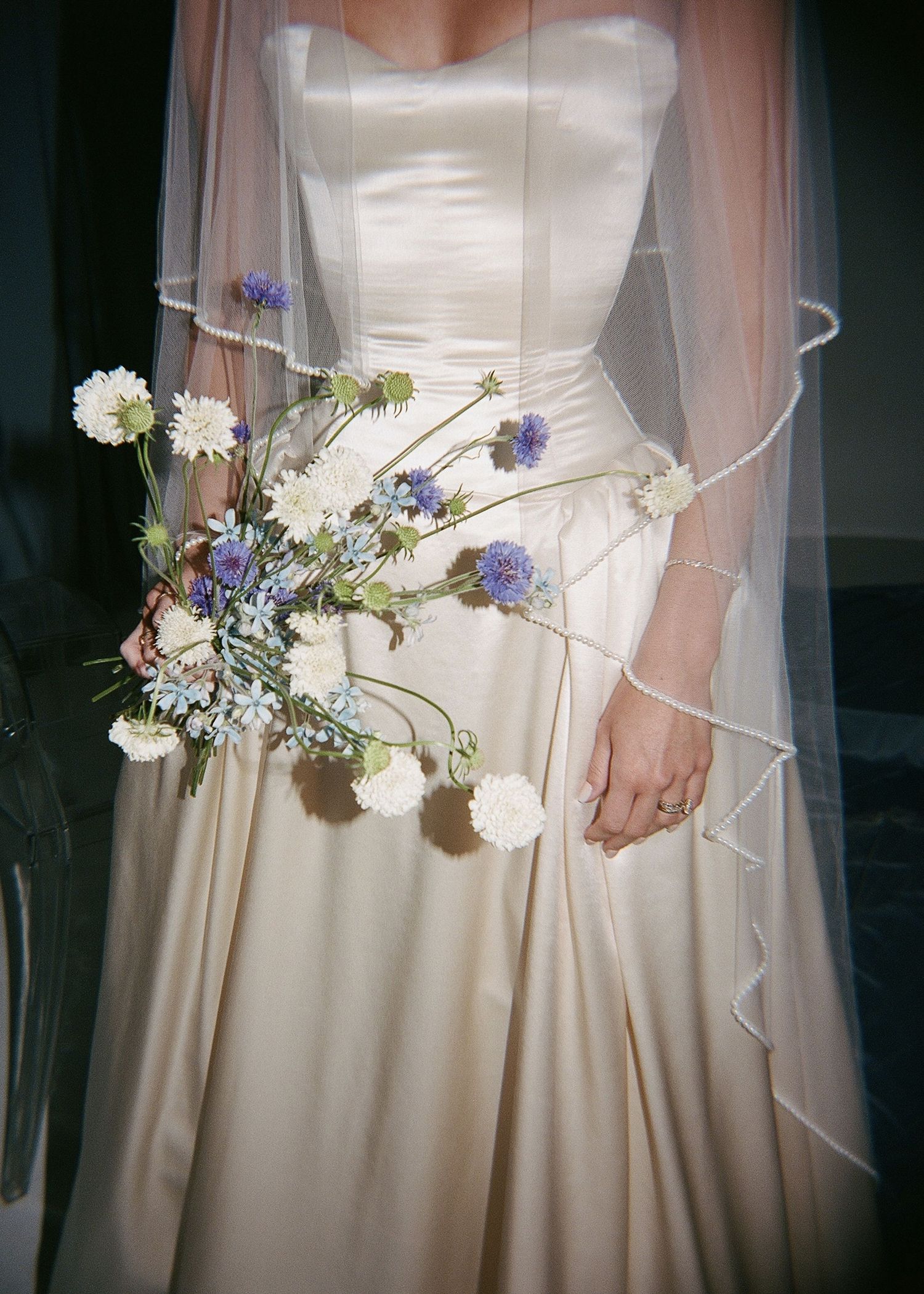Bride in strapless ivory satin ballgown holding a wildflower bouquet with white and purple blooms, wearing a sheer veil.