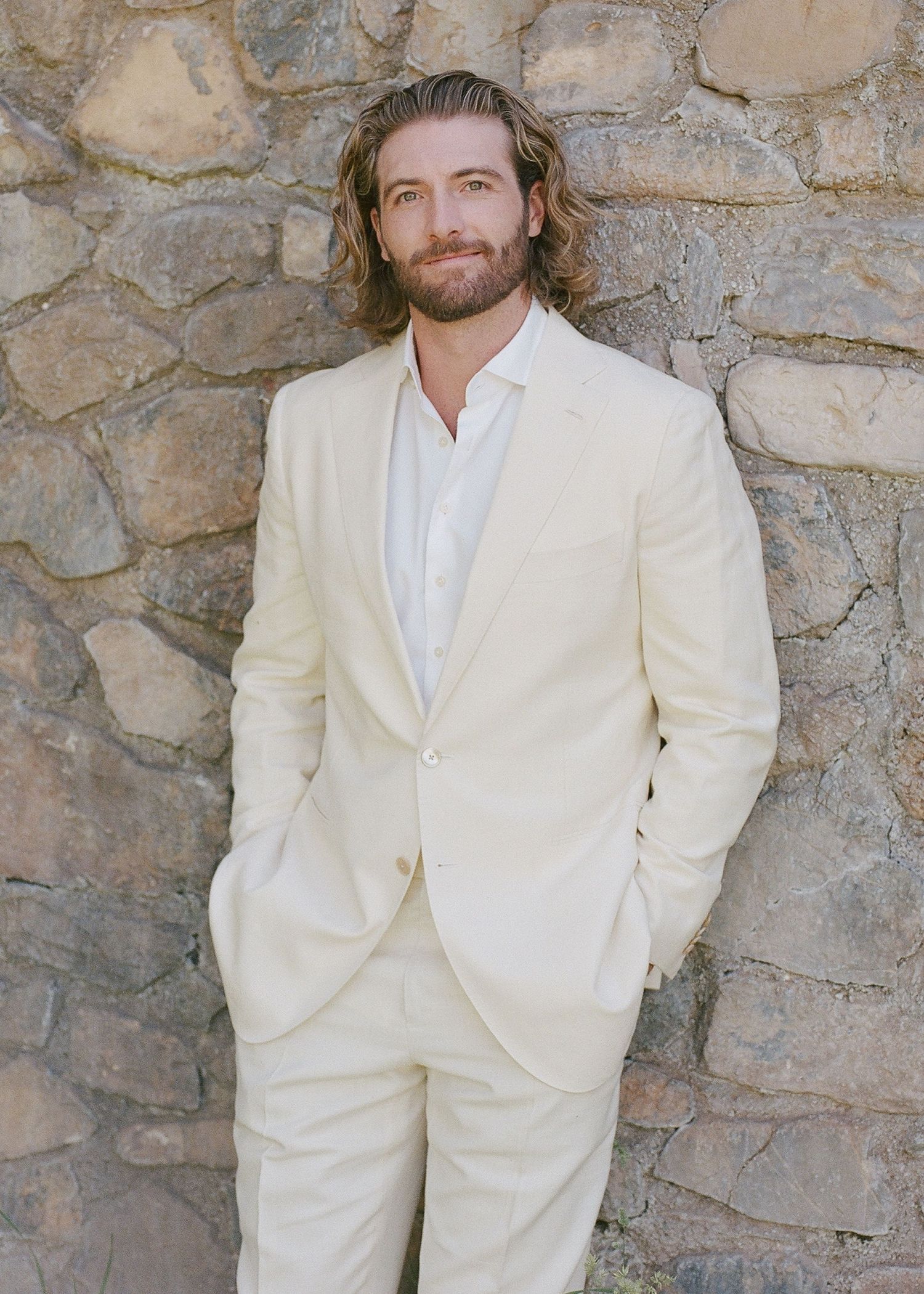 Man in cream suit with long hair and beard leaning against stone wall outdoors.