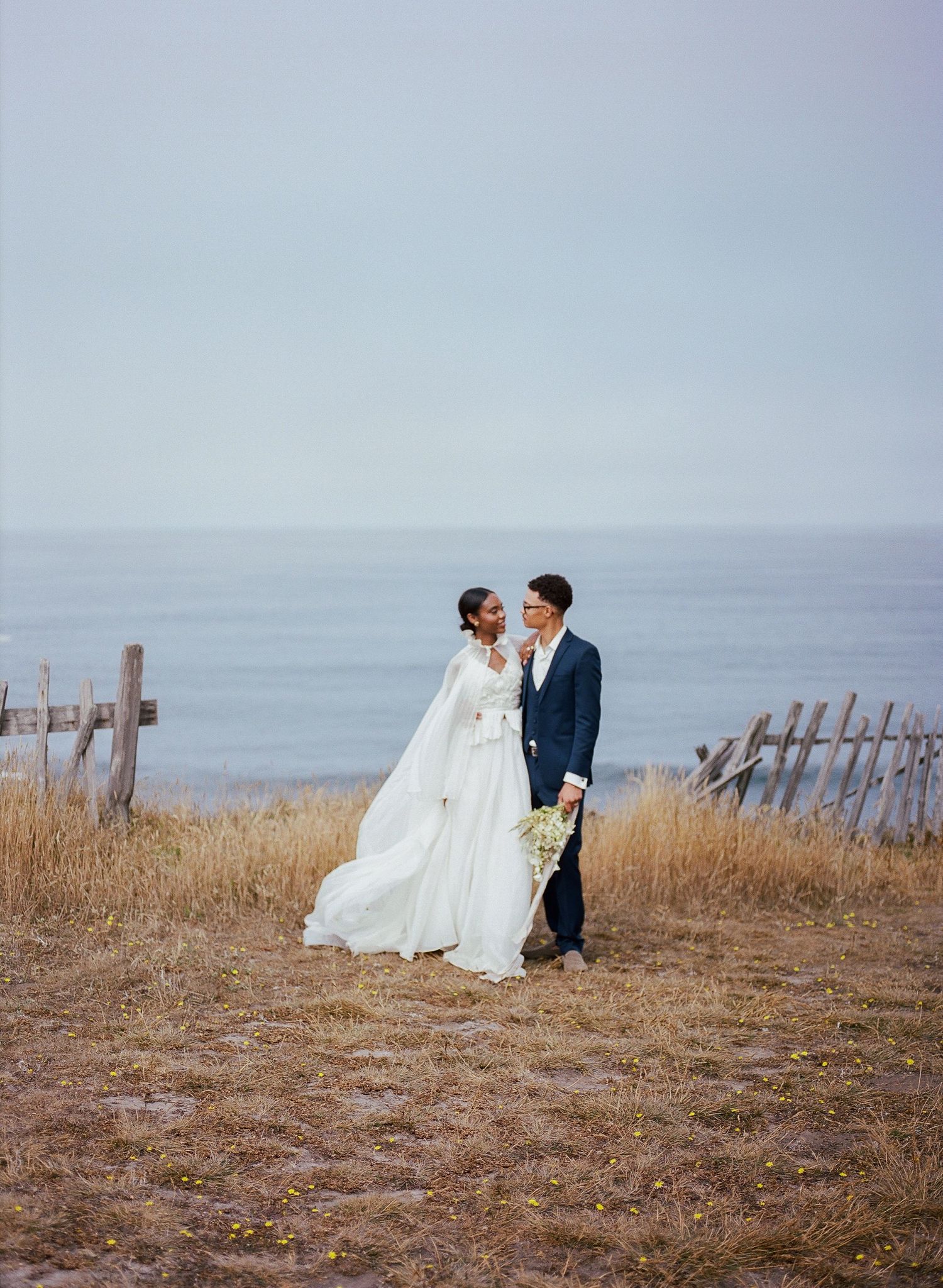 Bride and groom stand together in coastal field with ocean backdrop, she in white gown, he in navy suit.