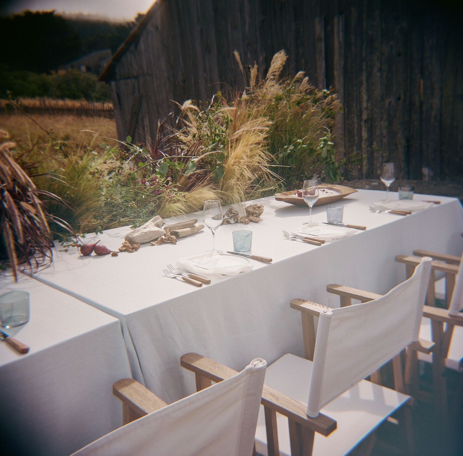 Elegant outdoor farm dinner table set with white linen, wildflower centerpieces, and wooden chairs against a rustic barn backdrop.