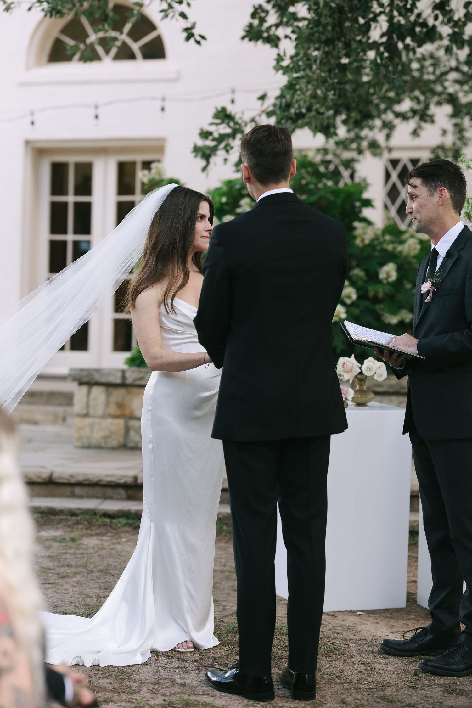 Intimate outdoor wedding ceremony with couple exchanging vows in front of white building.