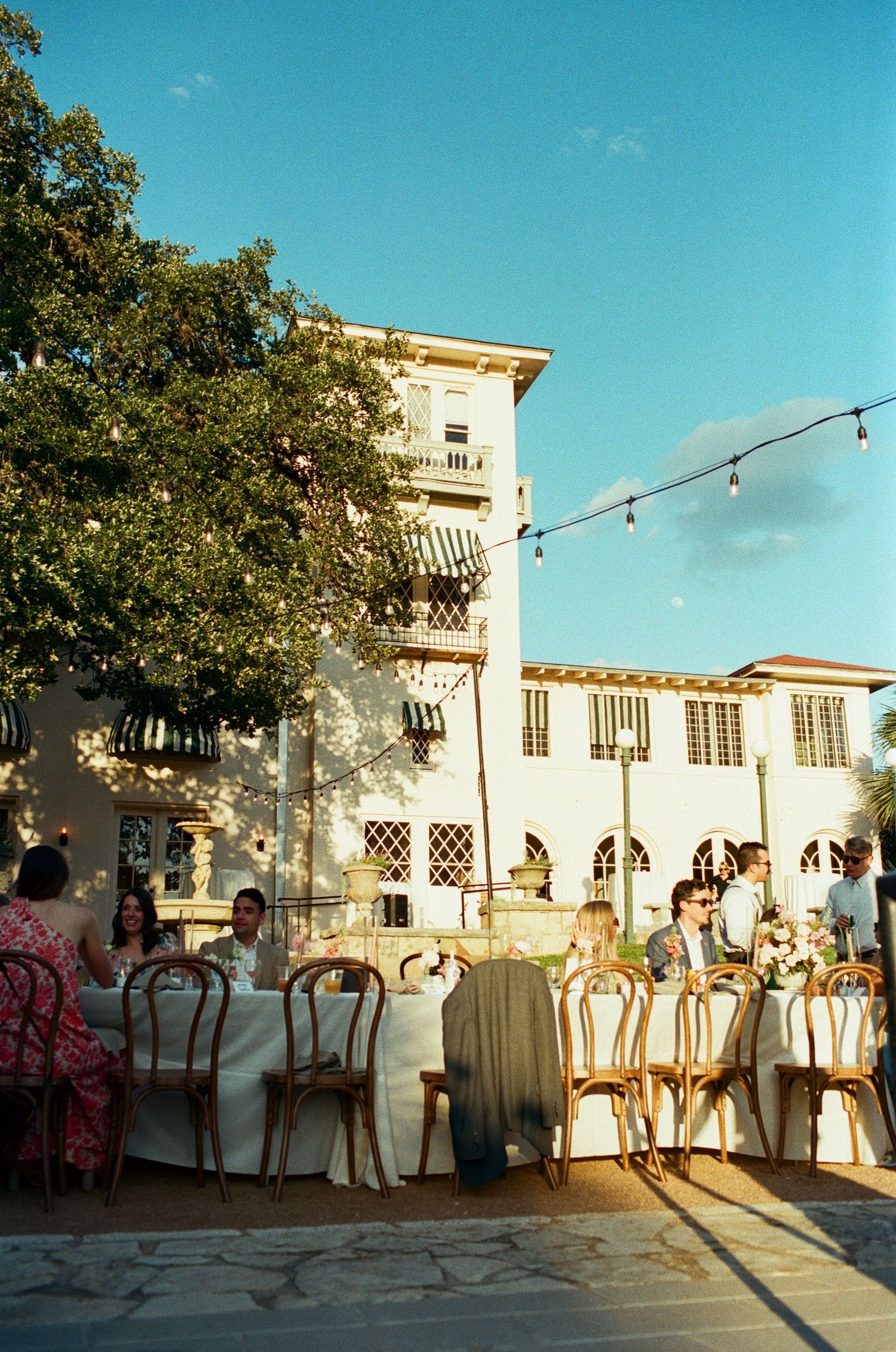 Wedding reception setup outside a white Spanish-style building with string lights at sunset.