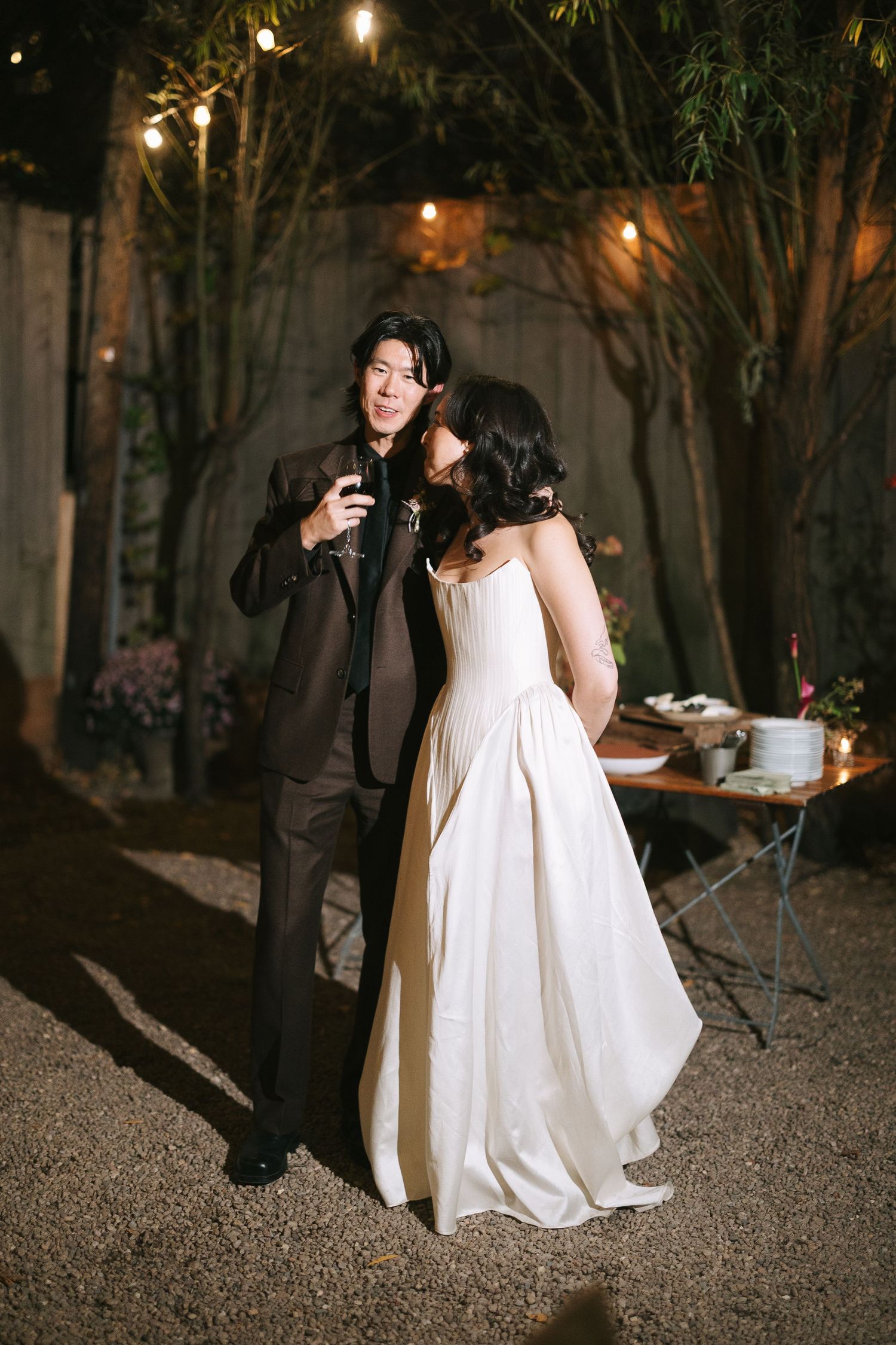 A couple shares a candid moment at their outdoor wedding reception under twinkling lights.