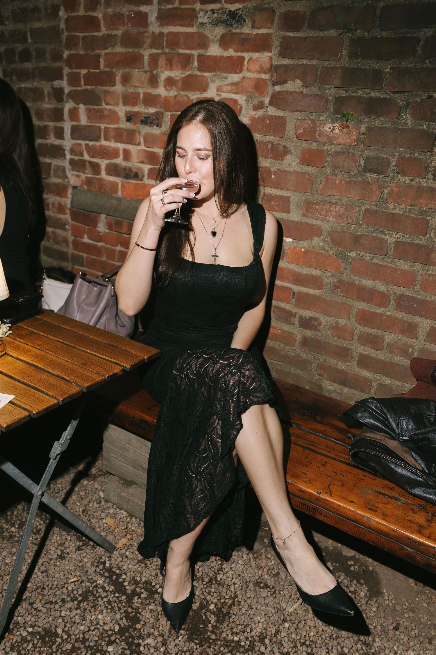 A woman in a black lace dress sits on a wooden bench enjoying a drink.