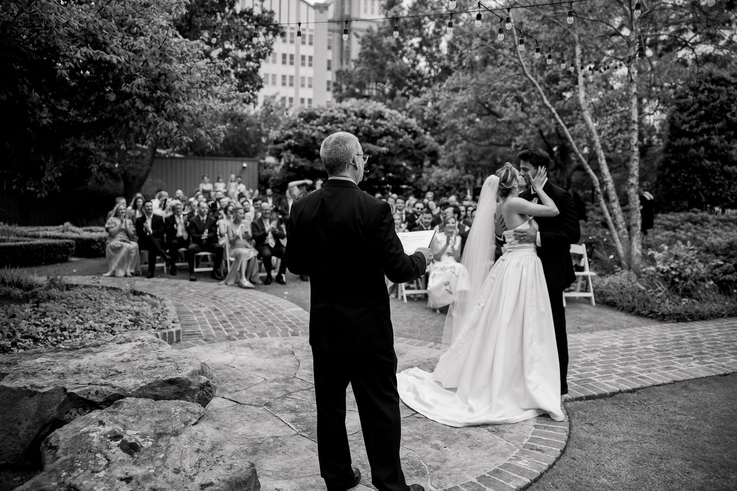 Outdoor wedding ceremony setup with guests gathered on a brick pathway surrounded by greenery.
