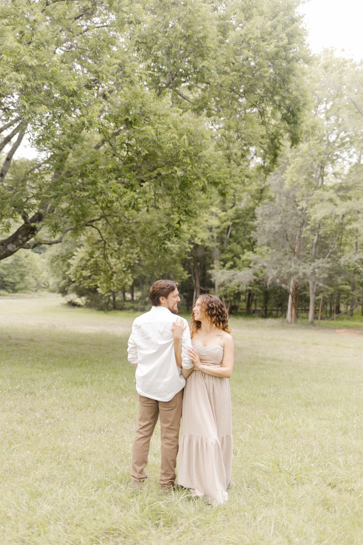 A couple in light-colored outfits walk together through a grassy field with trees in the background in Durham, North Carolina.