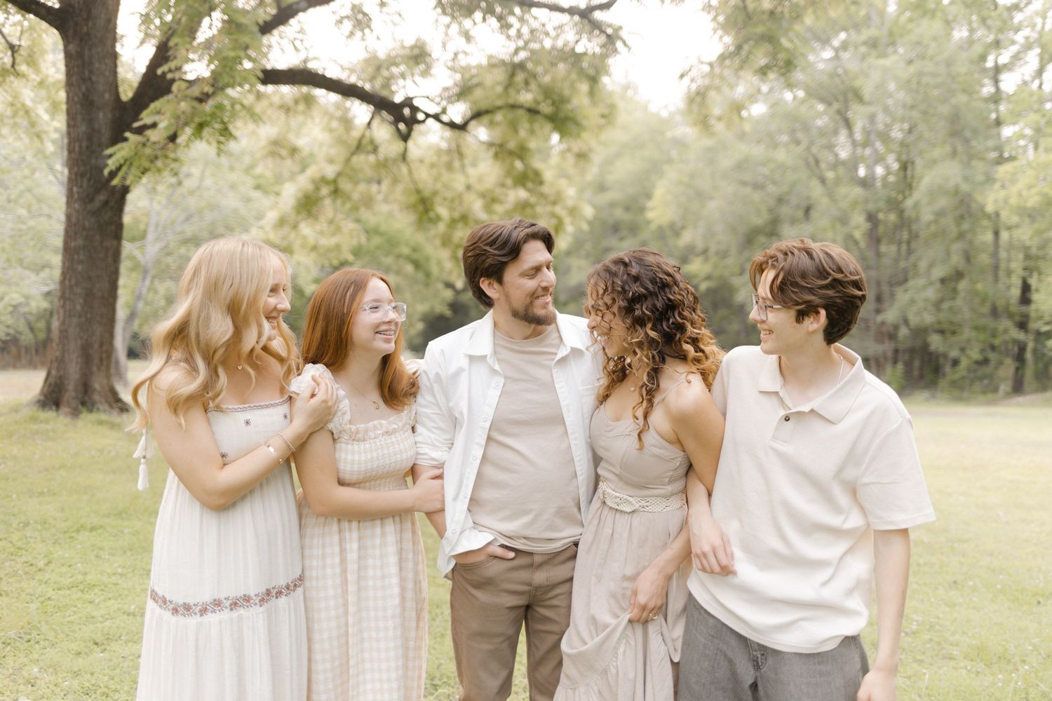 A group in light colored clothing laughs together while standing in a sunlit park setting during photoshoot with Kinsley Nicole Photography.