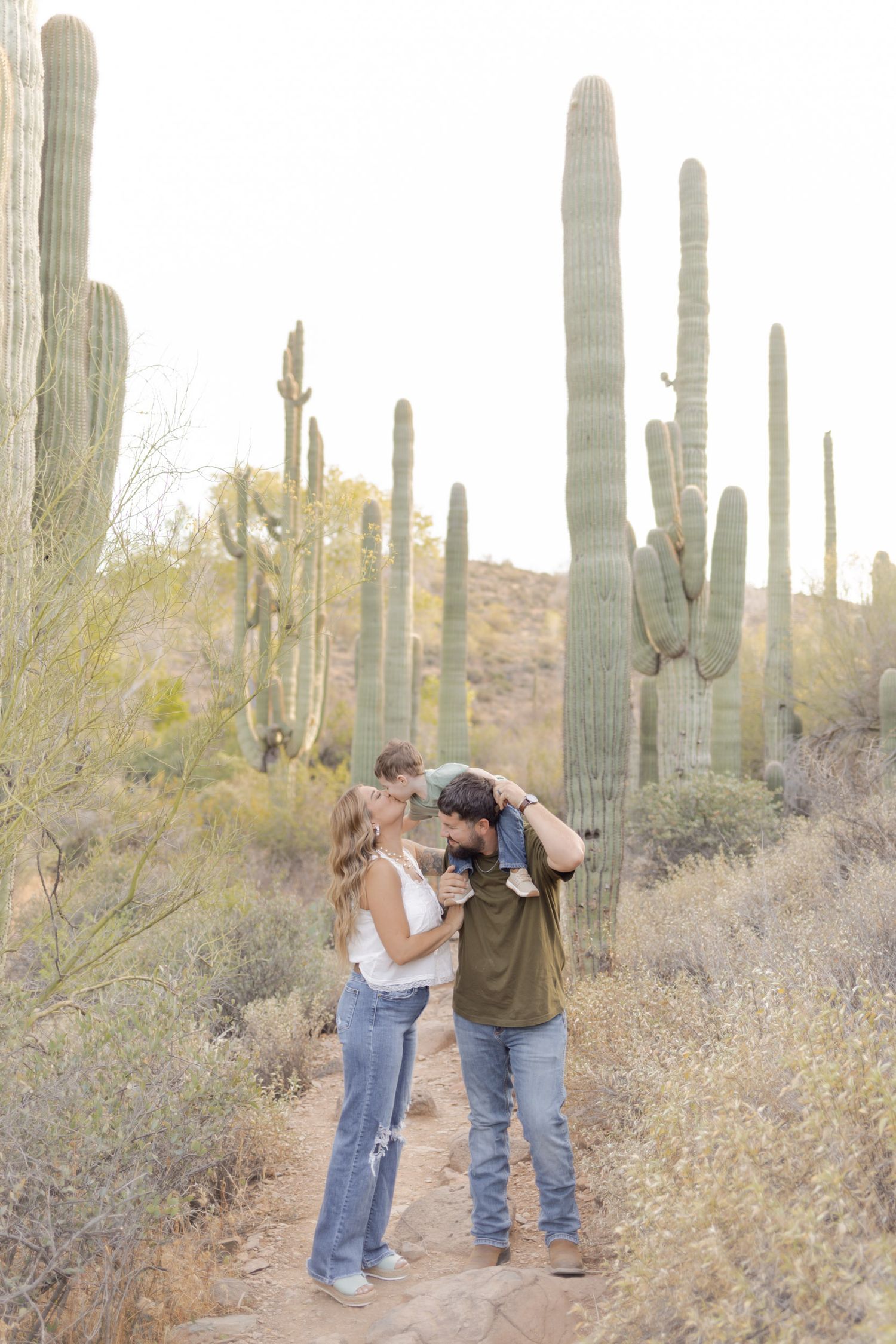 Couple sharing a kiss while holding toddler among tall saguaro cacti in Sonoran Desert.
