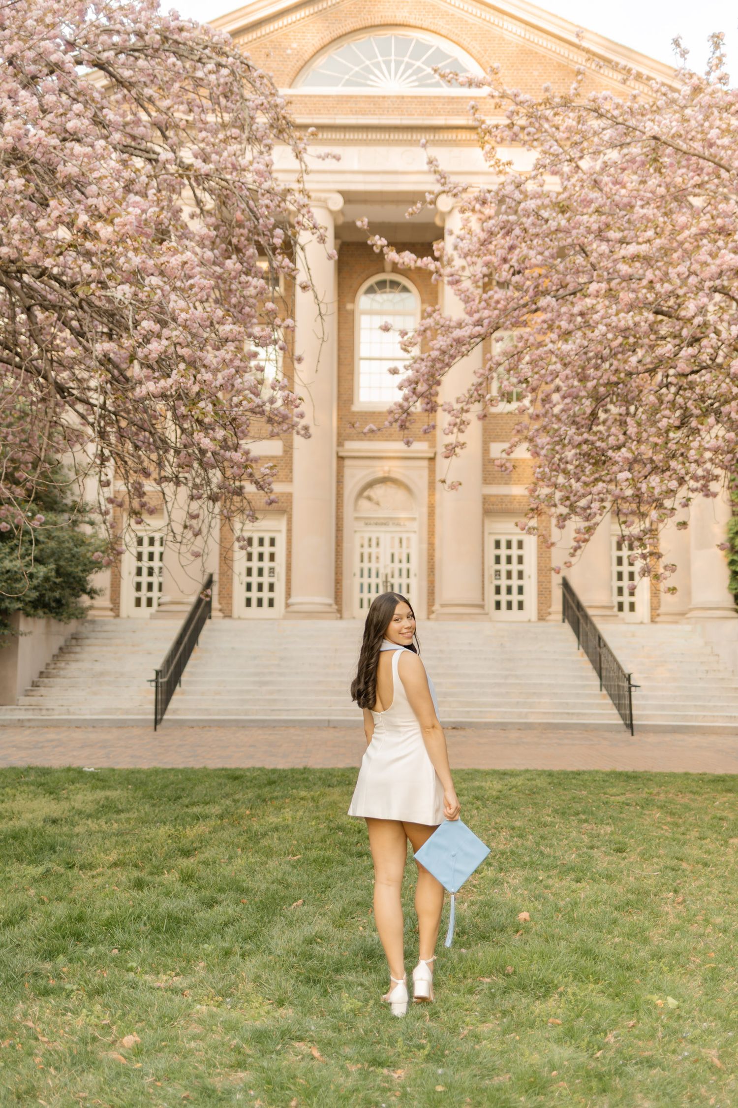 Young woman in white dress holding graduation cap poses before blooming cherry trees at a classic university building.