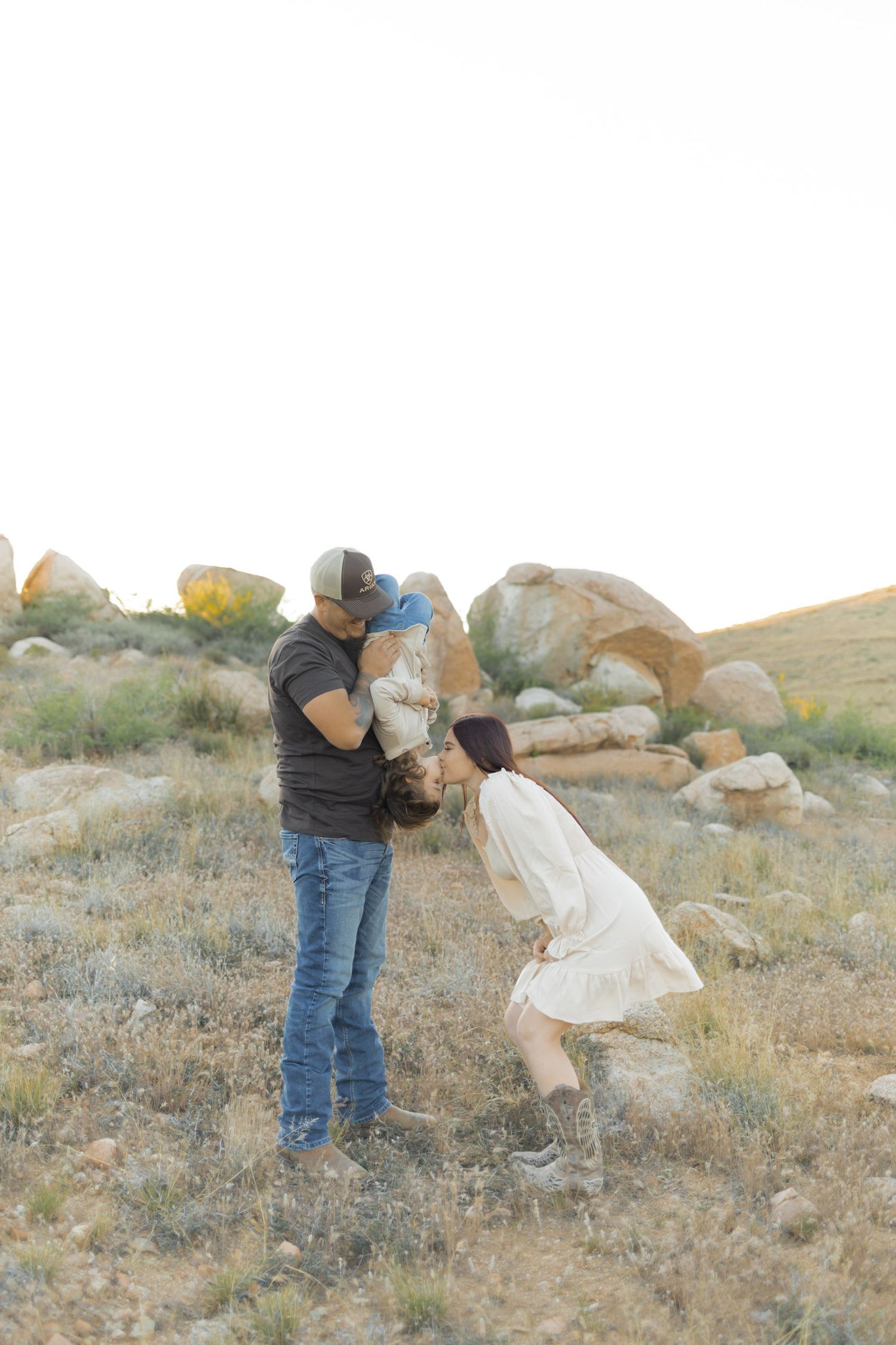 A family poses outdoors in a desert landscape, with a man lifting a baby while a woman in a white dress leans in to kiss.