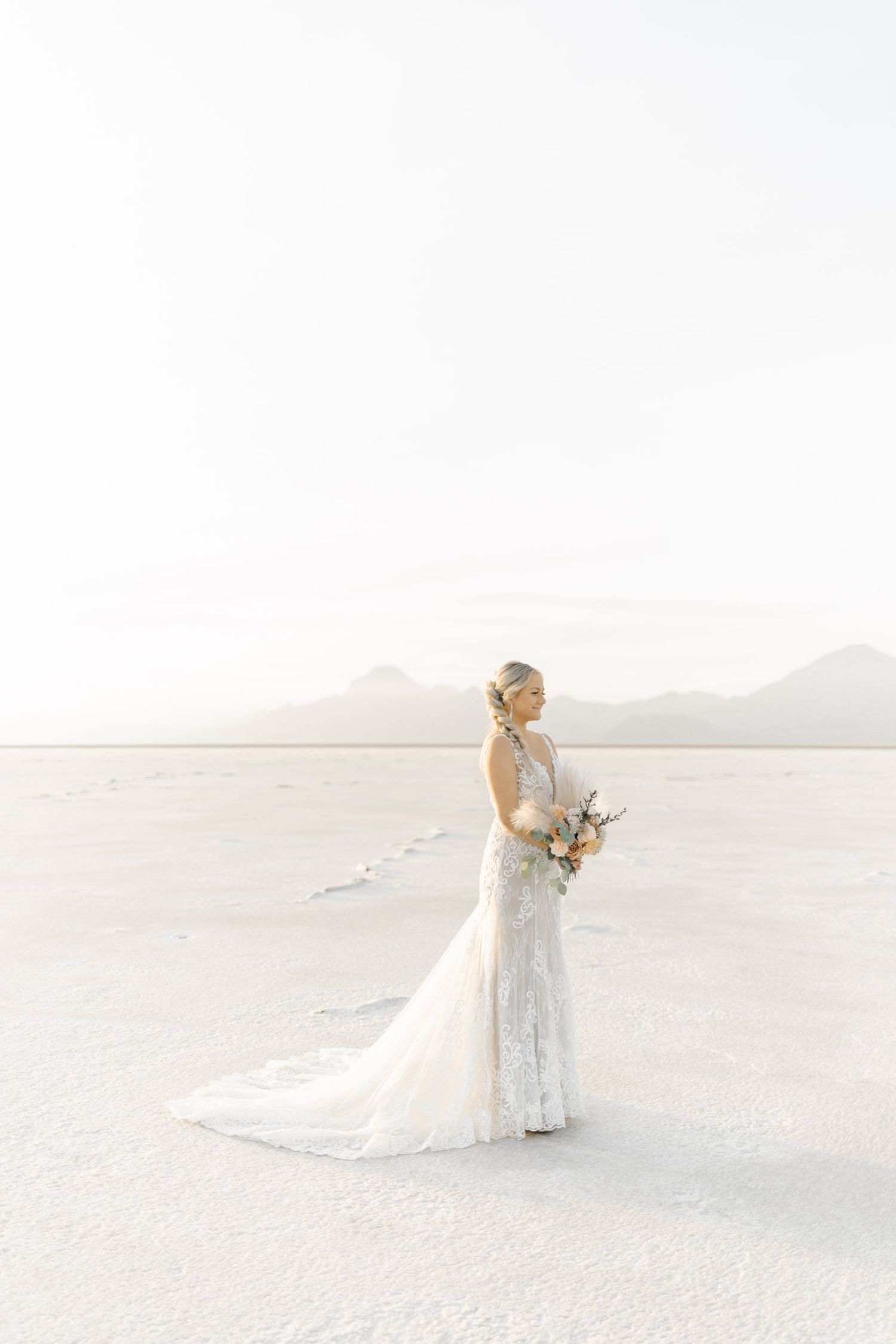 A bride in a flowing white lace gown stands elegantly on a stark white salt flat landscape during a desert wedding photoshoot.