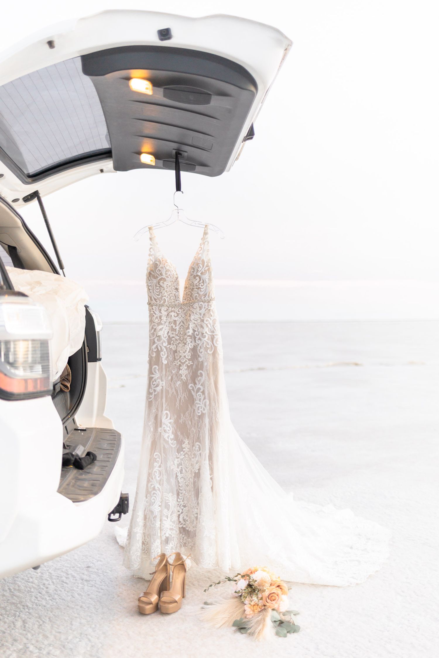 A delicate lace wedding dress hangs elegantly with nude heels and floral bouquet beneath a beauty light.