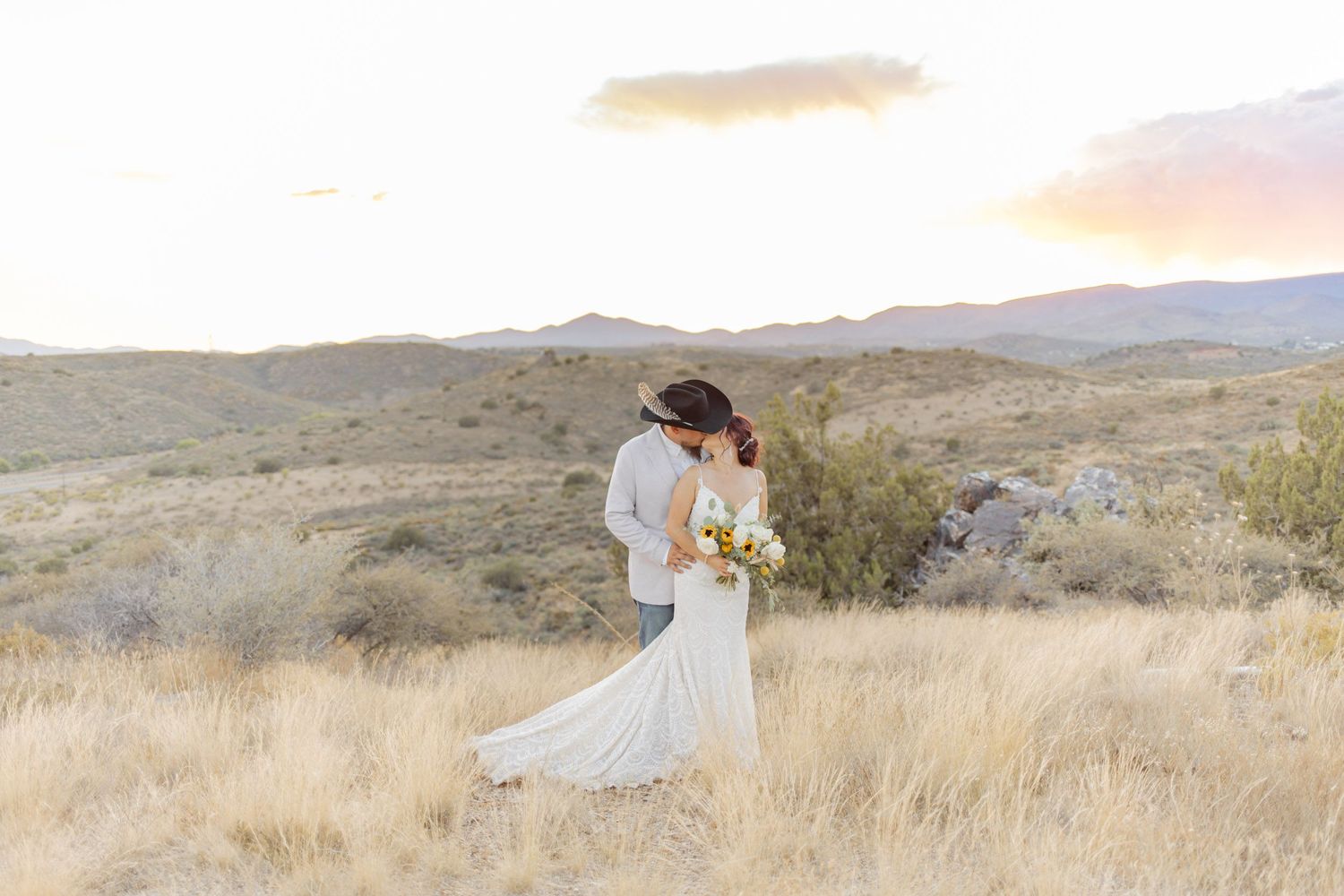 Bride and groom embracing in golden desert field at sunset with mountain landscape backdrop.