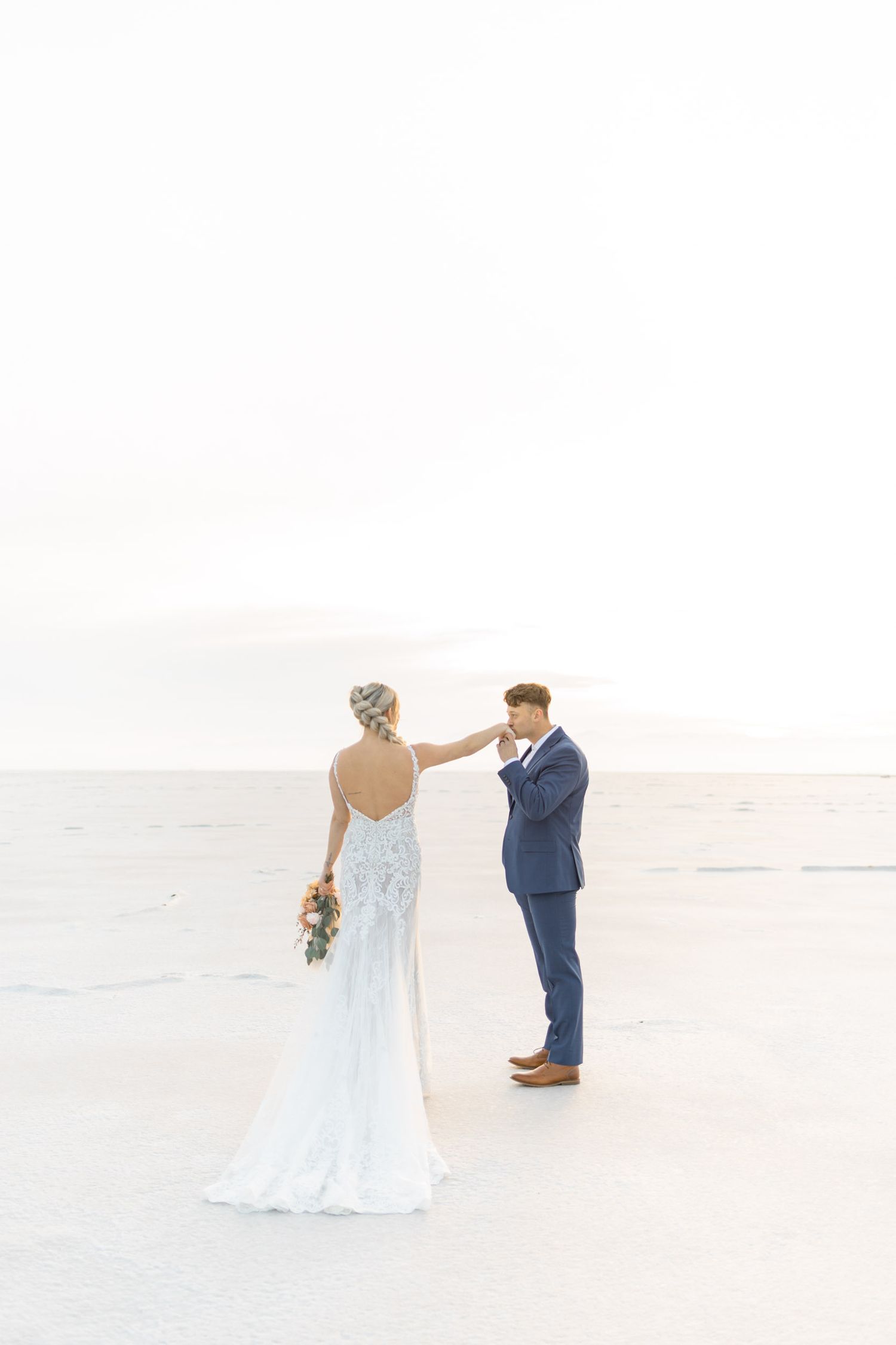Newlyweds walk hand in hand across a bright white salt flat landscape with mountains in the distance.