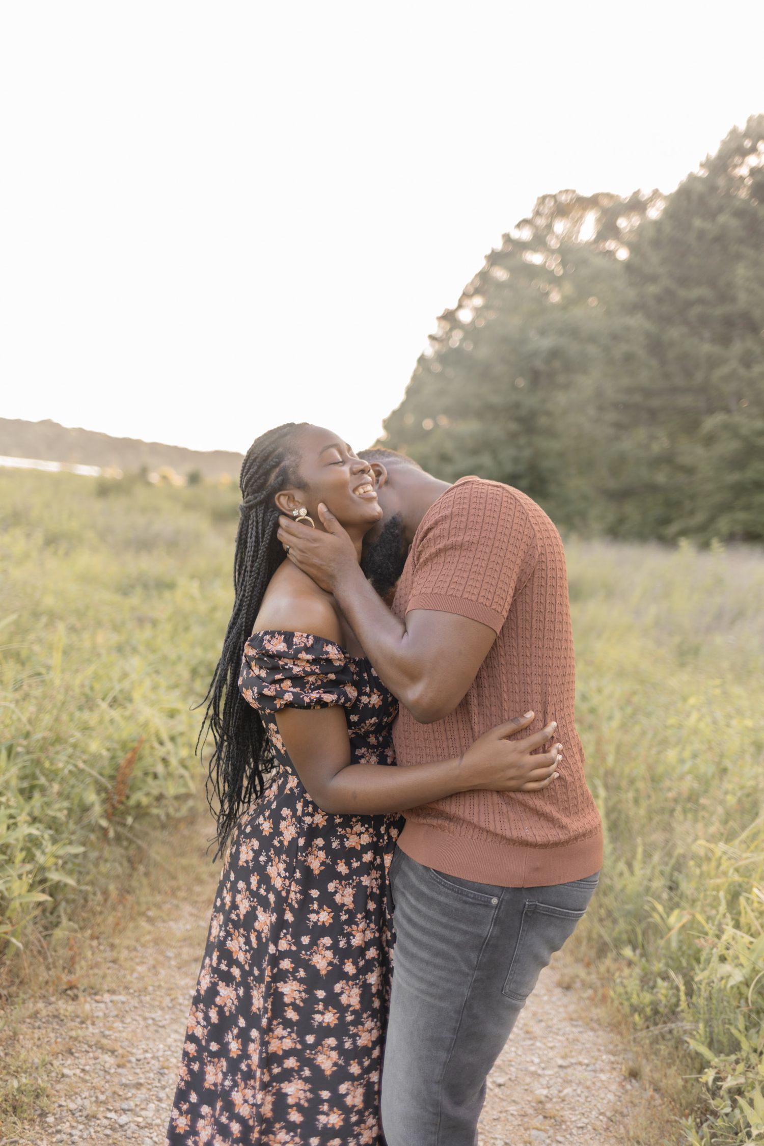 A joyful couple embraces and laughs together on a scenic path surrounded by lush greenery at golden hour.