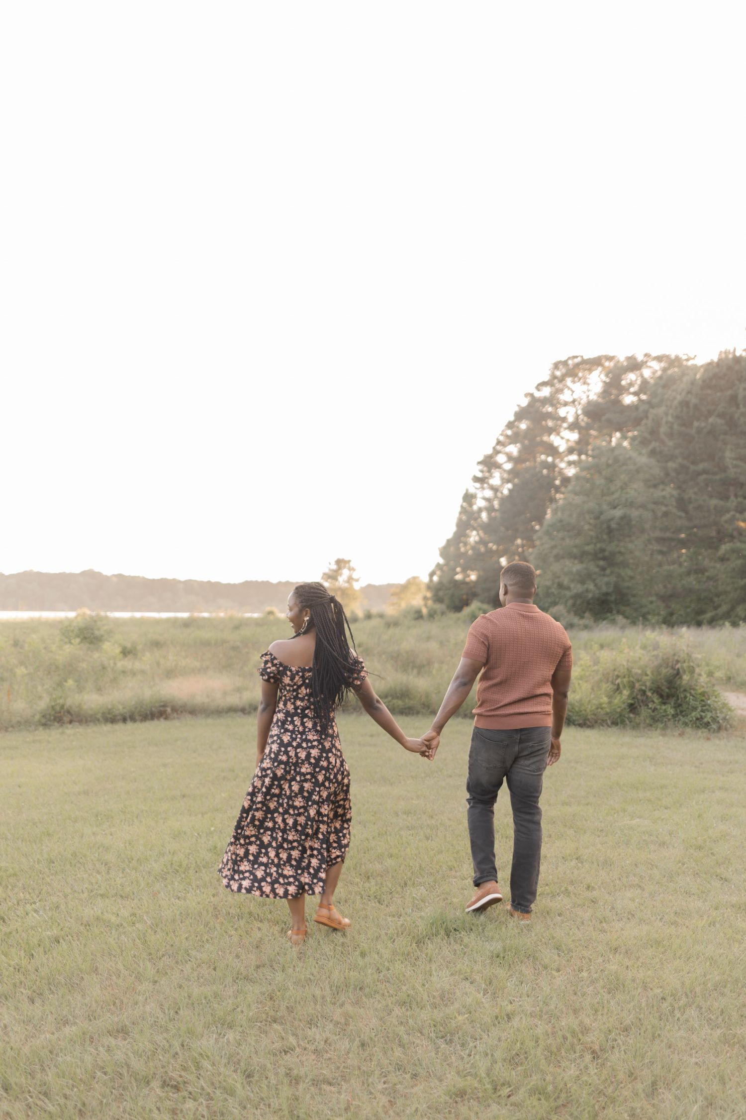 A couple holds hands walking through a green field at sunset with trees and water in the background.