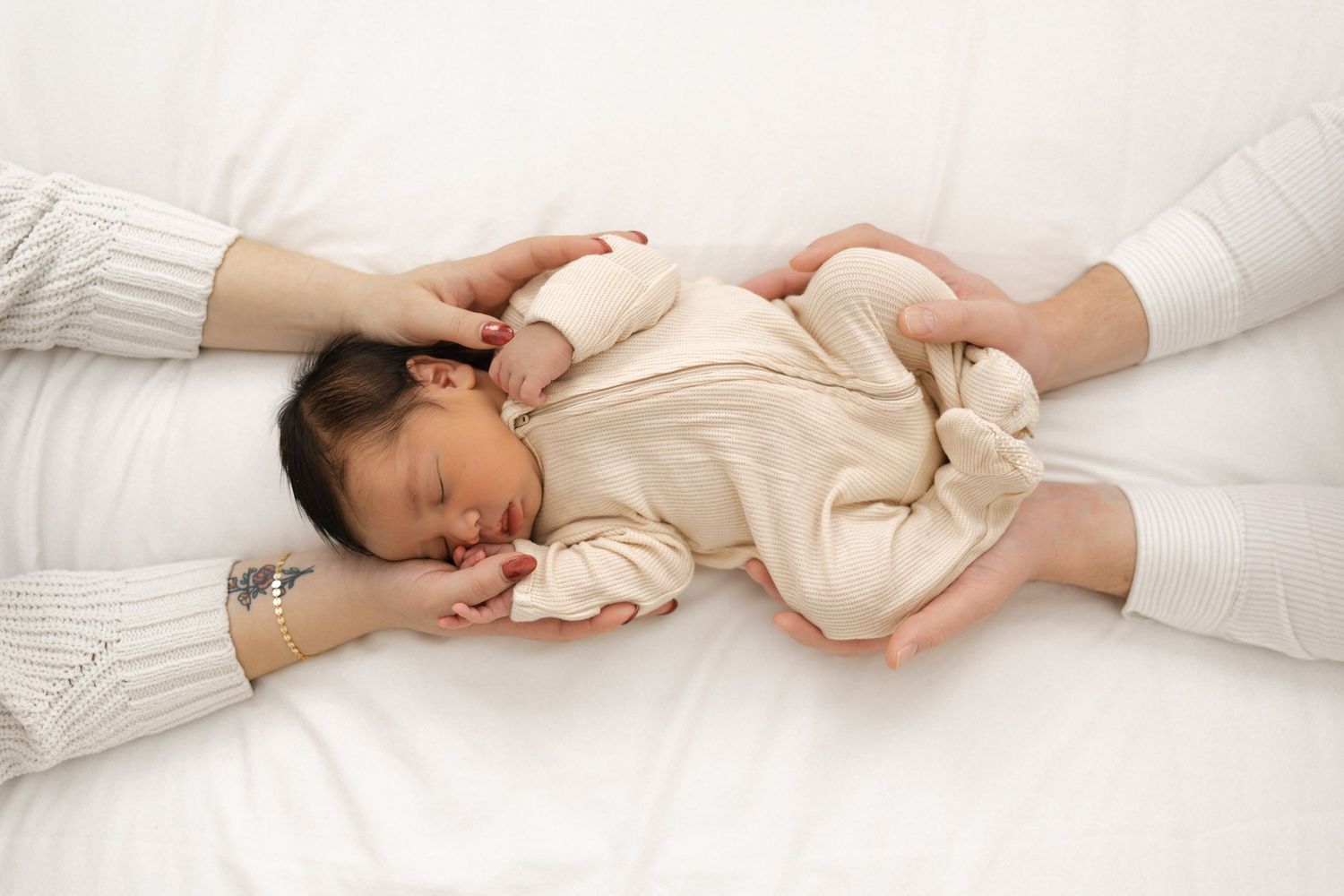 Sleeping newborn in beige onesie cradled by parents hands on white background.