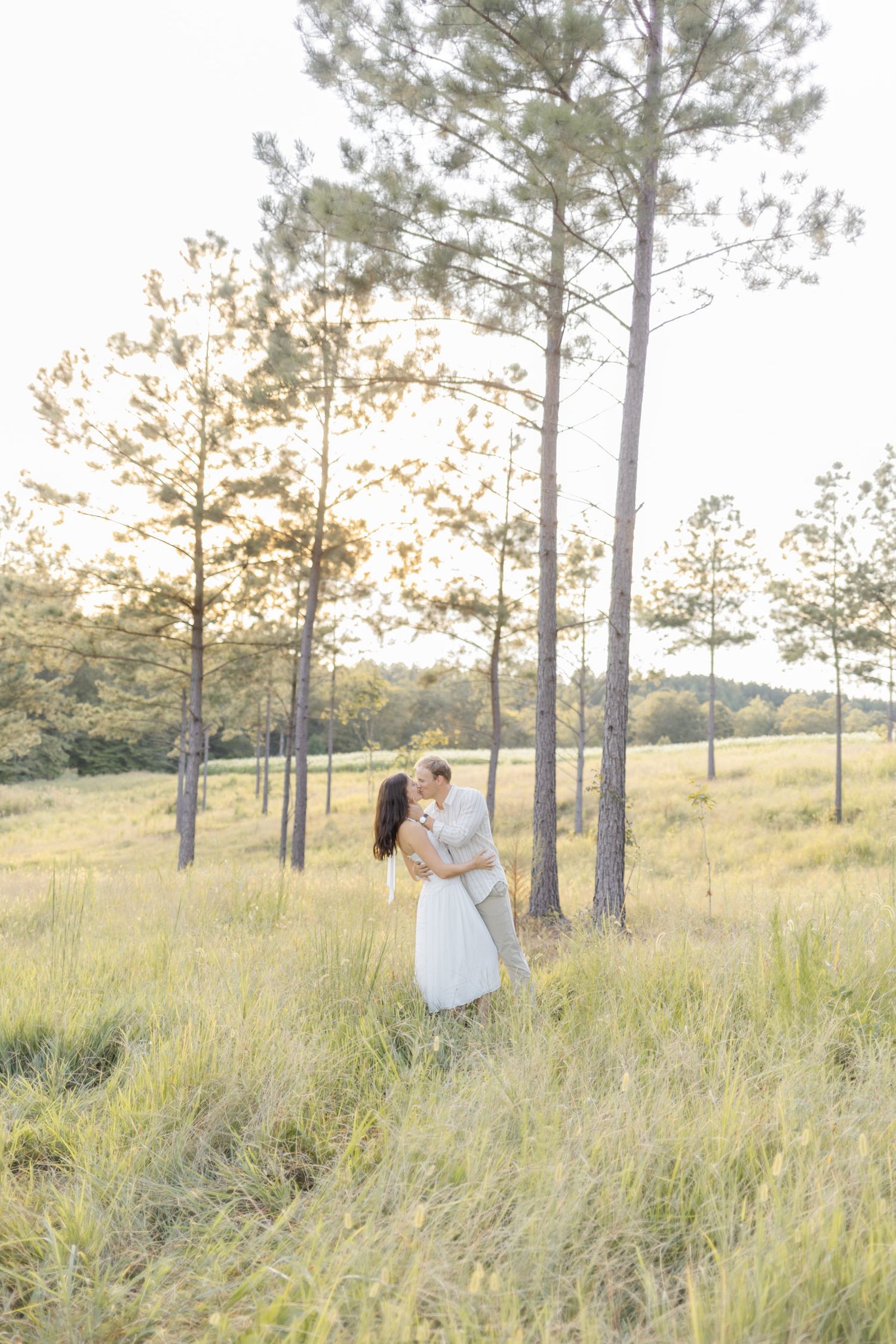 Intimate couple sharing tender moments in a sunlit field surrounded by pine trees making a light, airy, creamy photo.