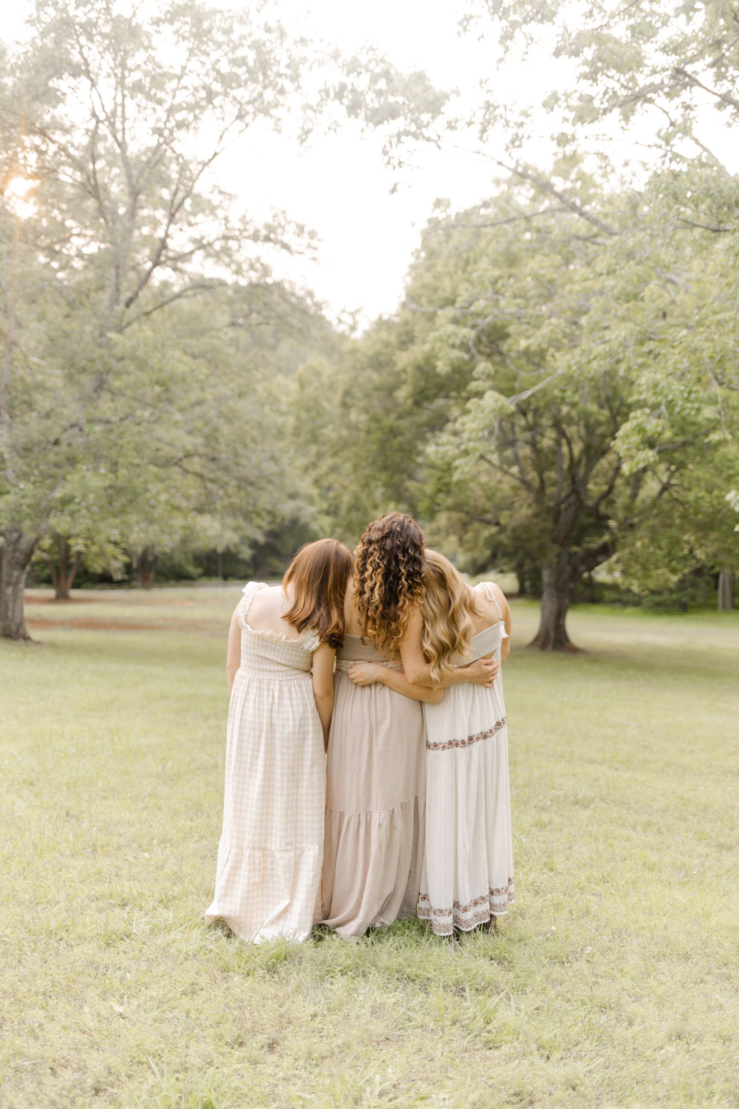 Friends in coordinating cream dresses stand together in an embrace against a backdrop of green trees in Durham, North Carolina.