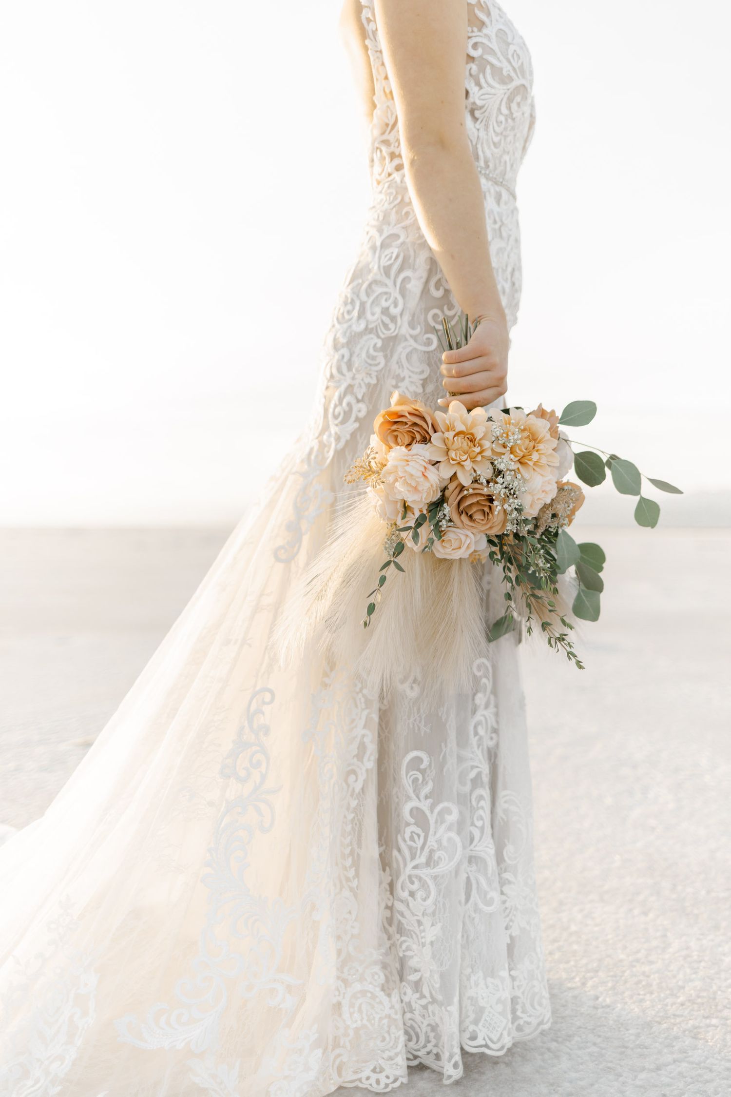 Close-up of a delicate lace wedding dress with a peach and cream floral bouquet featuring eucalyptus accents.