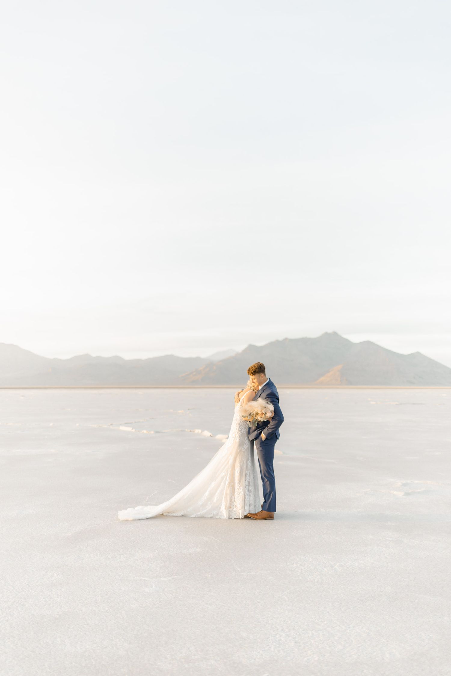 A romantic couple embraces on a vast salt flat landscape with mountains in the background at sunset.