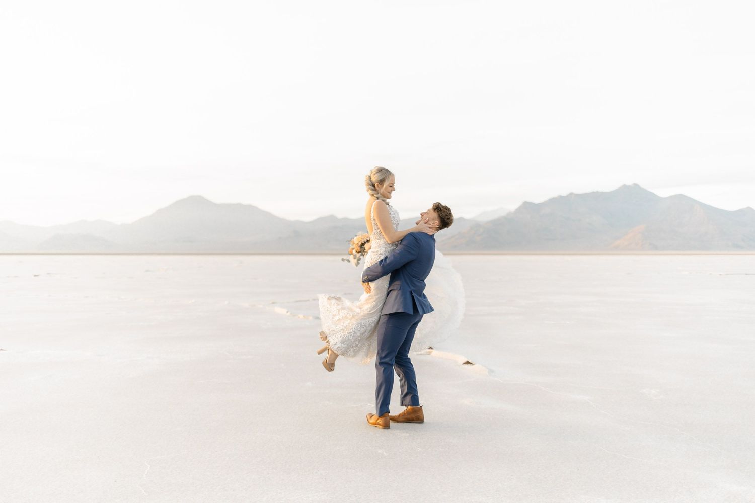 A couple performs a romantic lift during their wedding photoshoot on white salt flats.