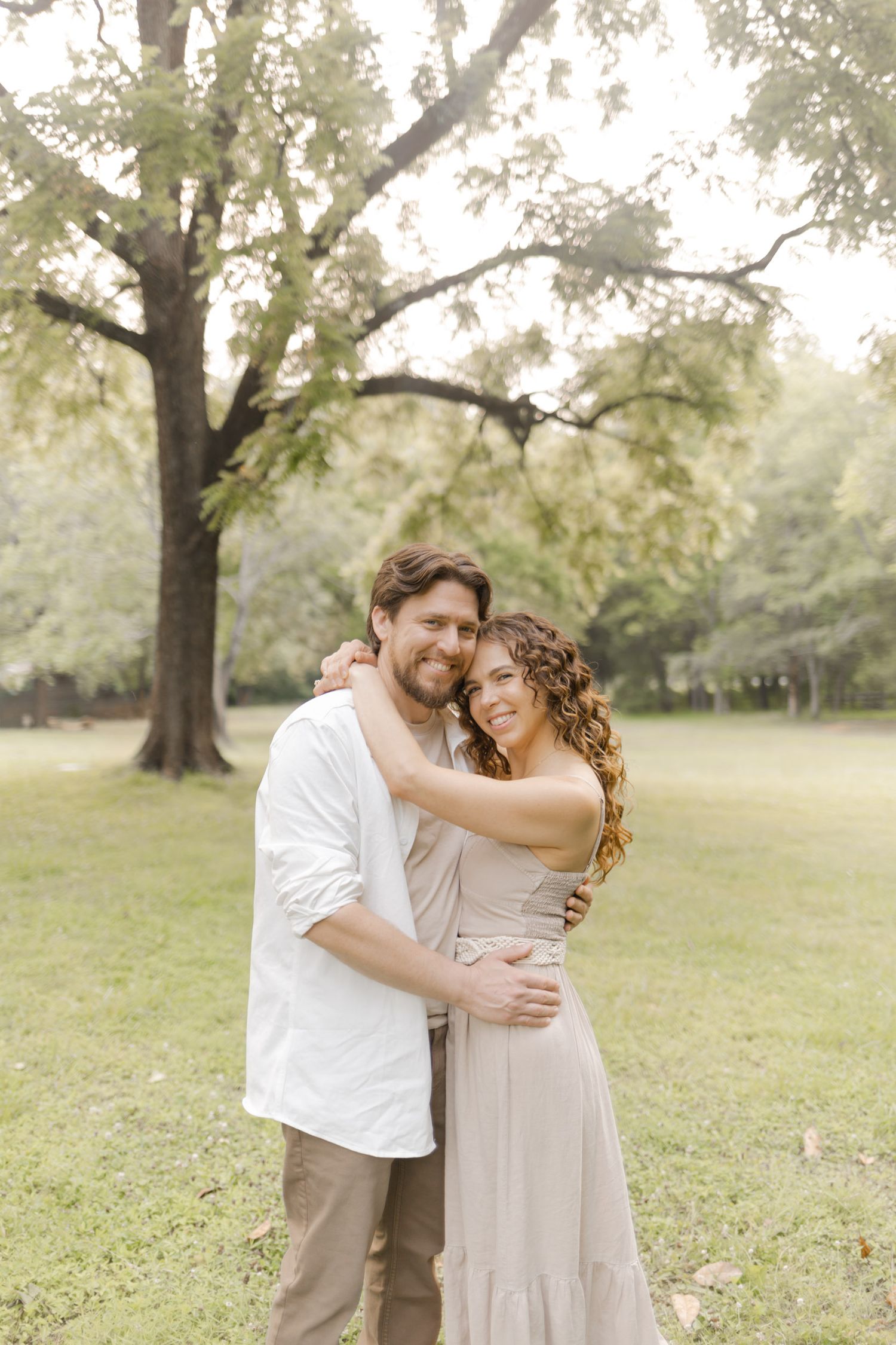 A romantic couple in light neutral clothing embraces in a sunlit park setting with mature trees in the background while doing a photoshoot.
