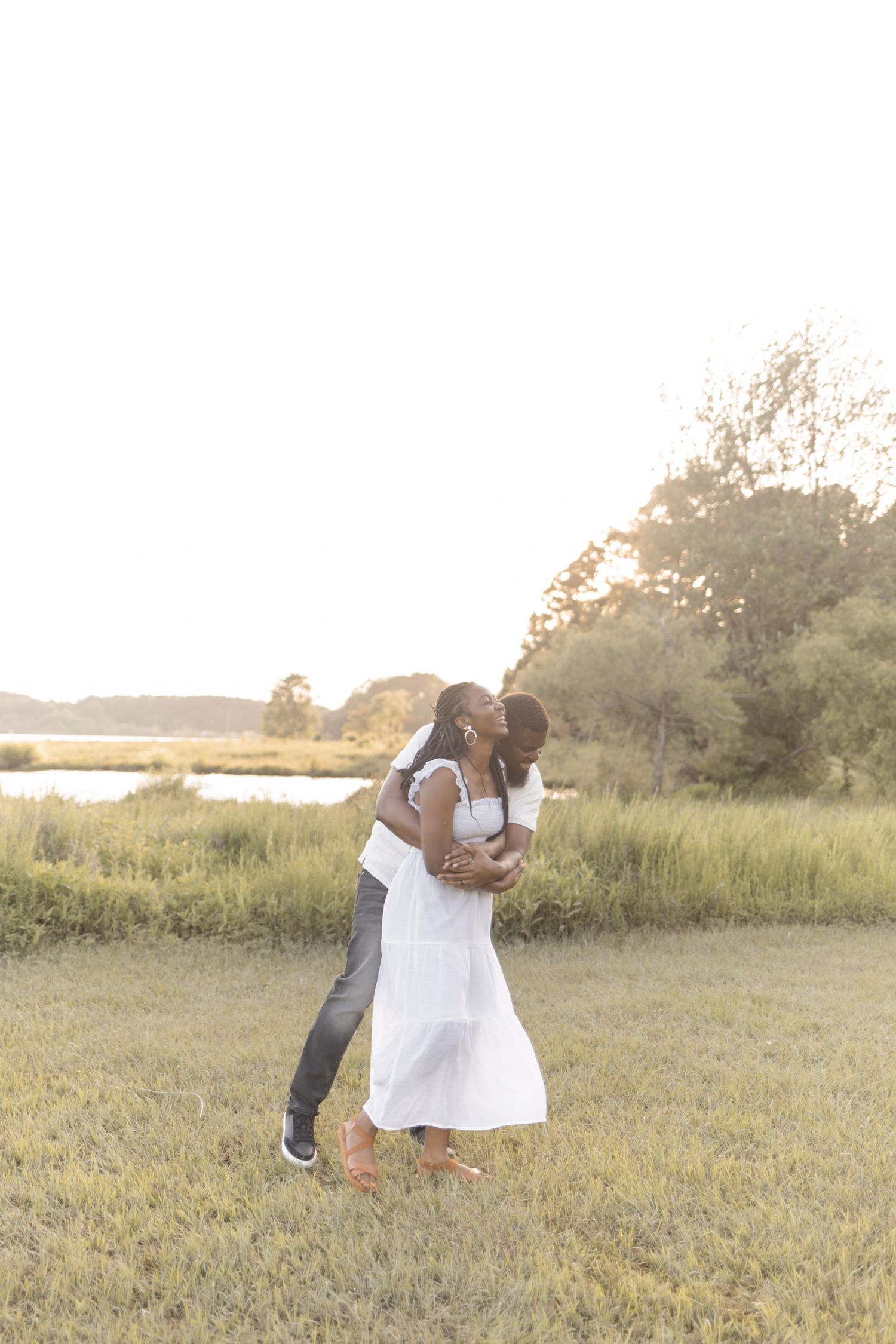 Couple sharing a romantic kiss outdoors at golden hour, woman in white dress, lush green field and water in background.