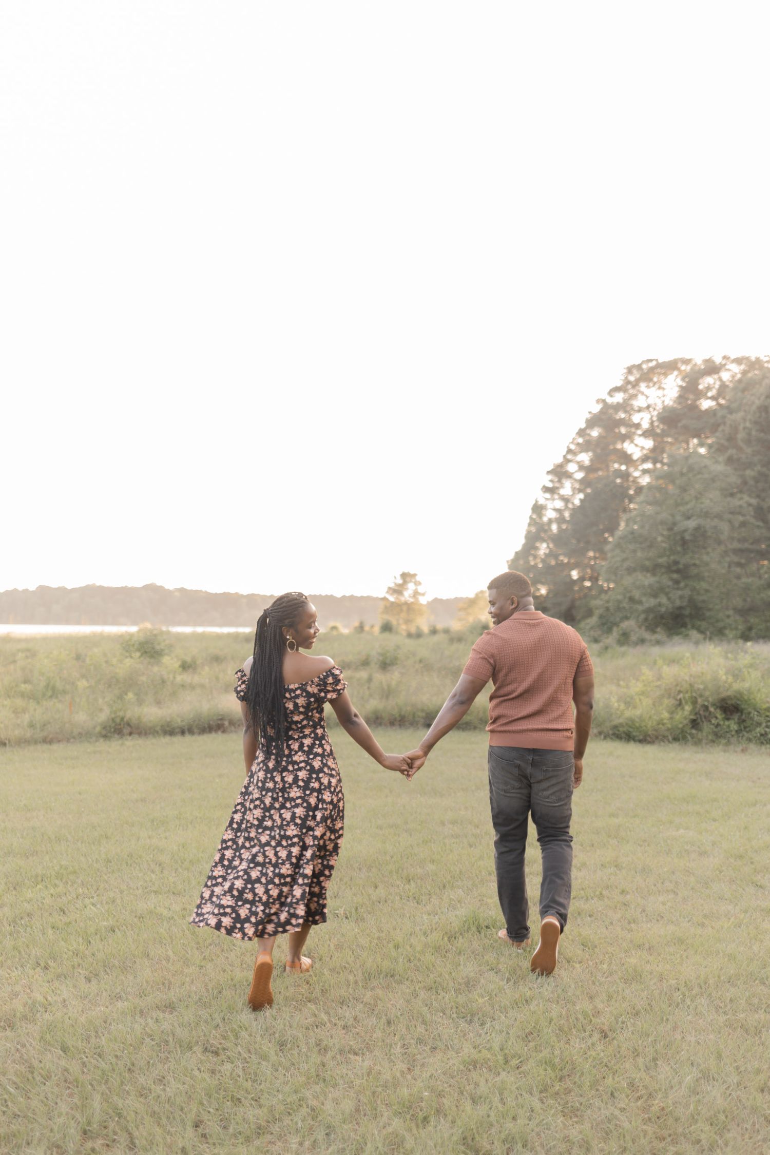 A couple holds hands walking through a green field at sunset, woman in floral dress, man in casual attire.