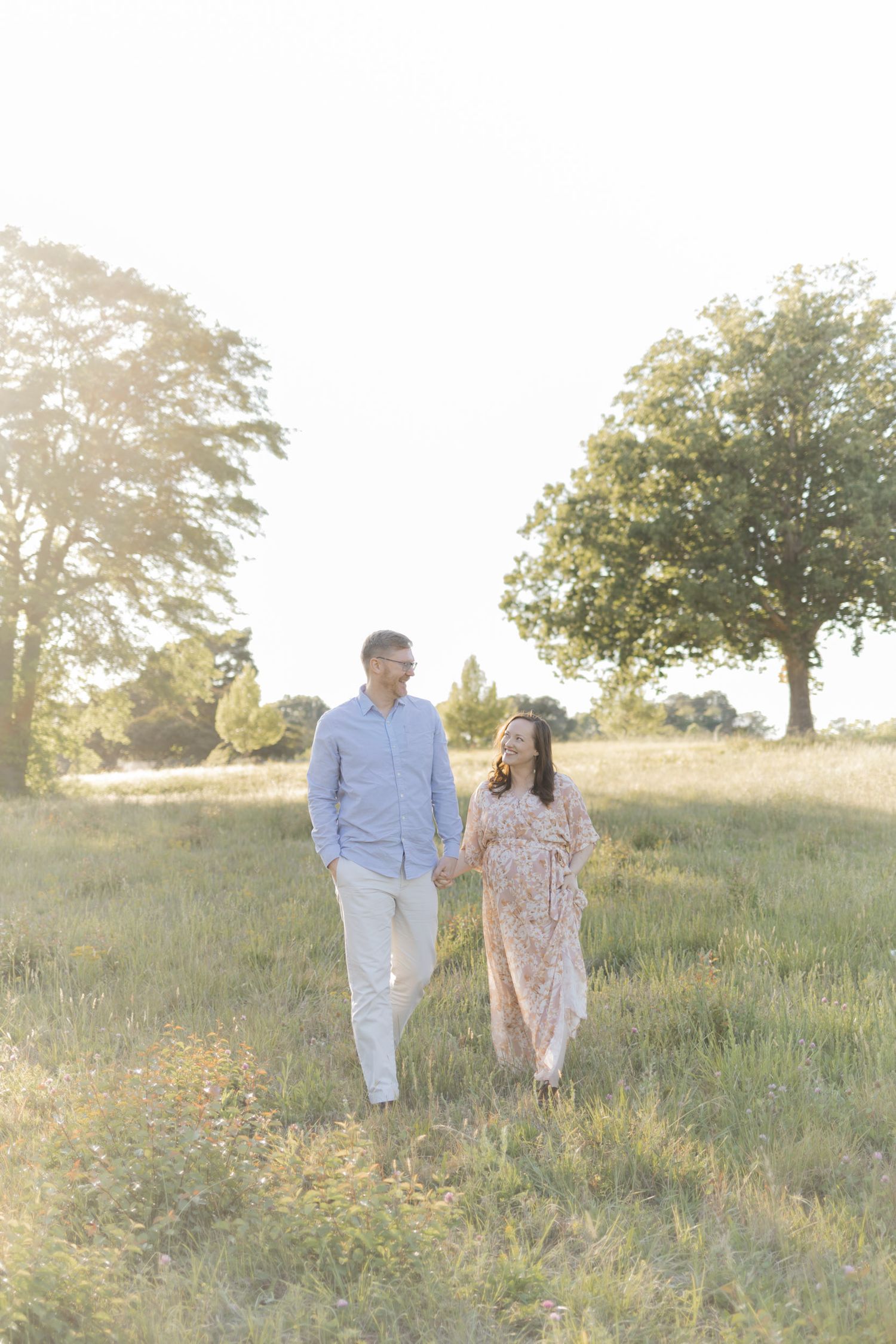Expecting couple holding hands walking through sunny green meadow, woman in floral maternity dress, golden hour light.