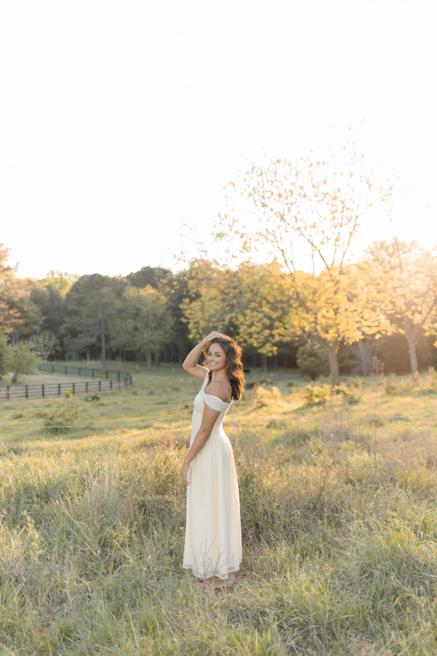 Young woman in cream maxi dress standing in sunlit field during golden hour.