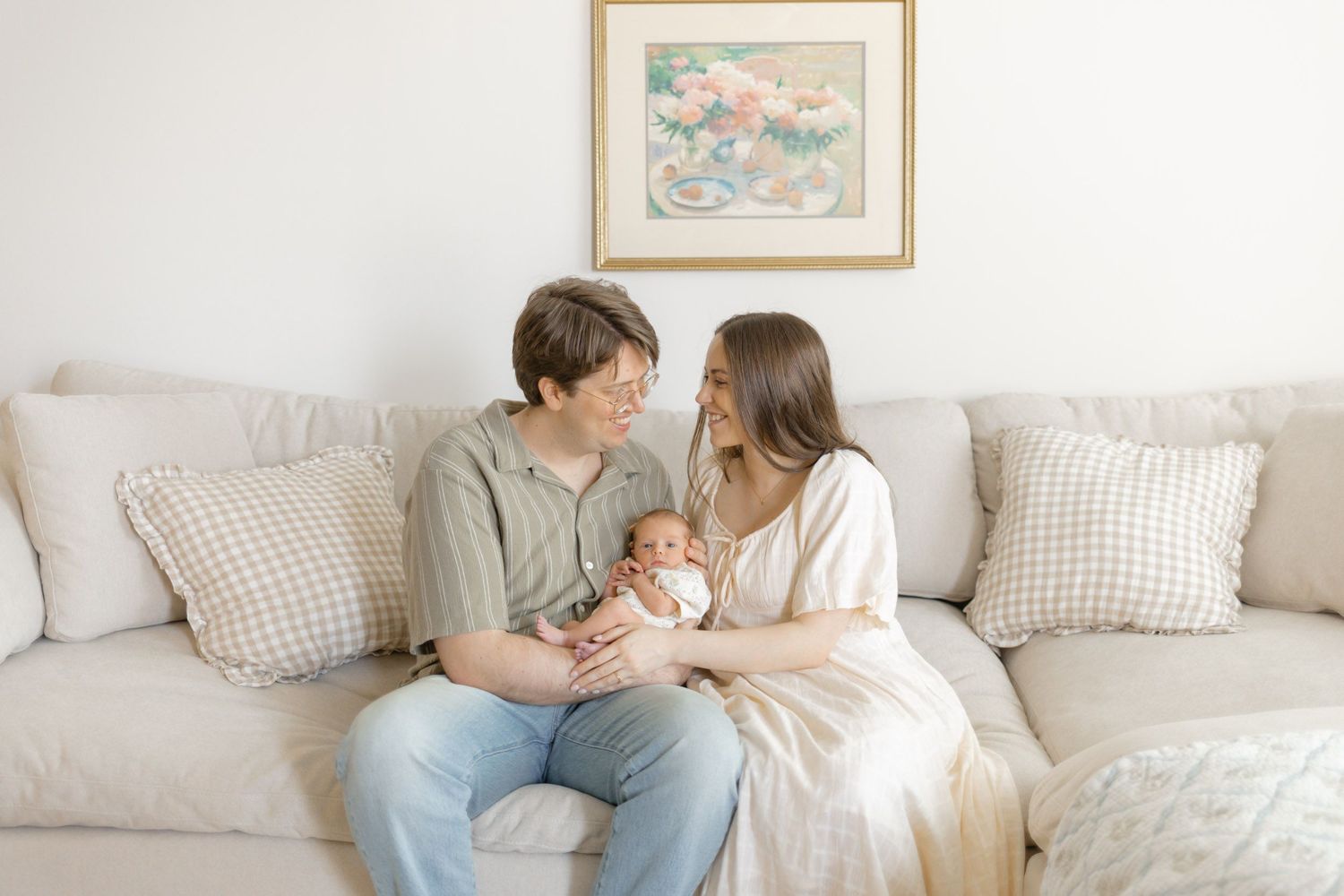 New parents sit on a beige couch smiling at their newborn baby in a bright, cozy living room.