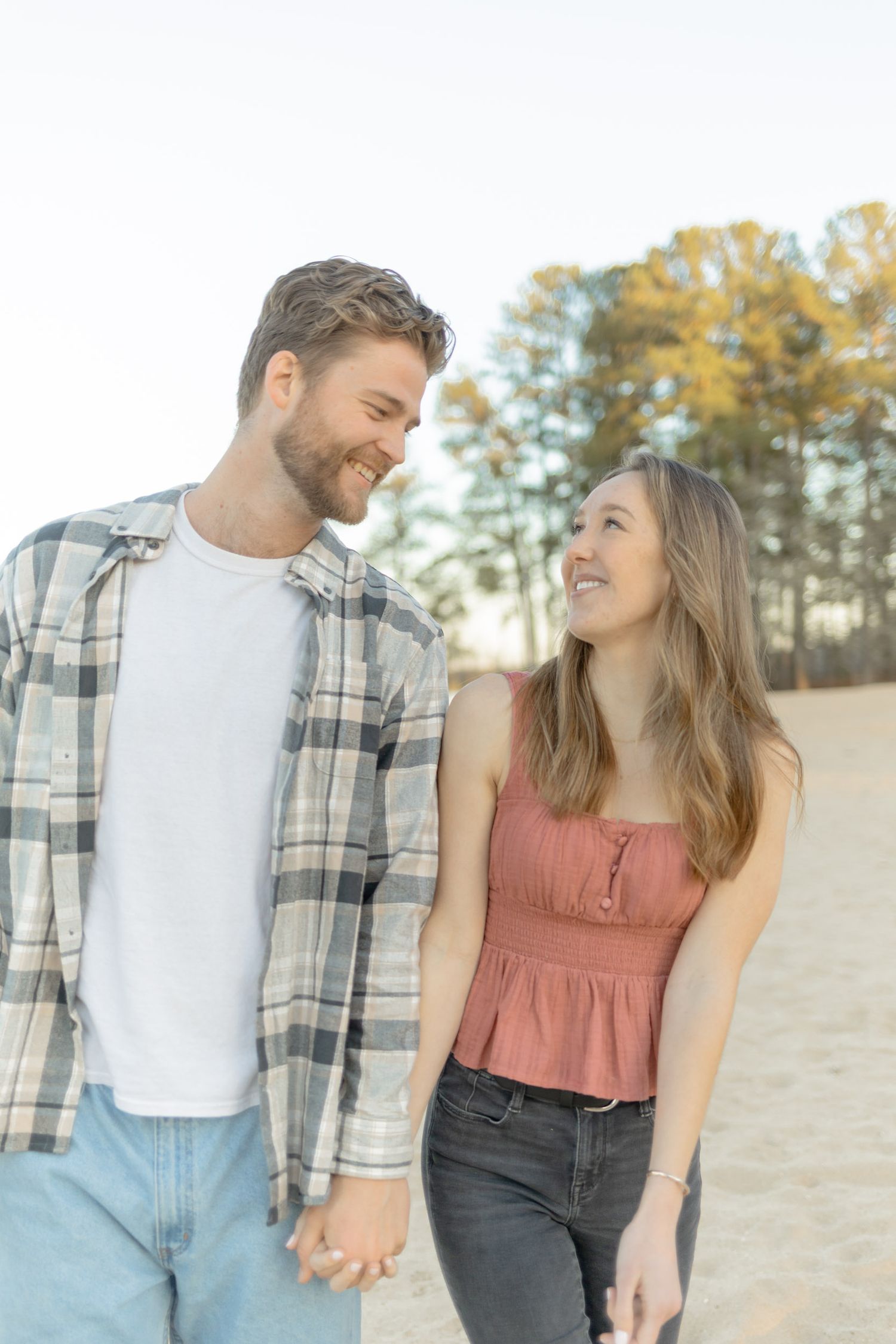 Couple holding hands and smiling while walking on sandy beach with pine trees in background in Raleigh, North Carolina.