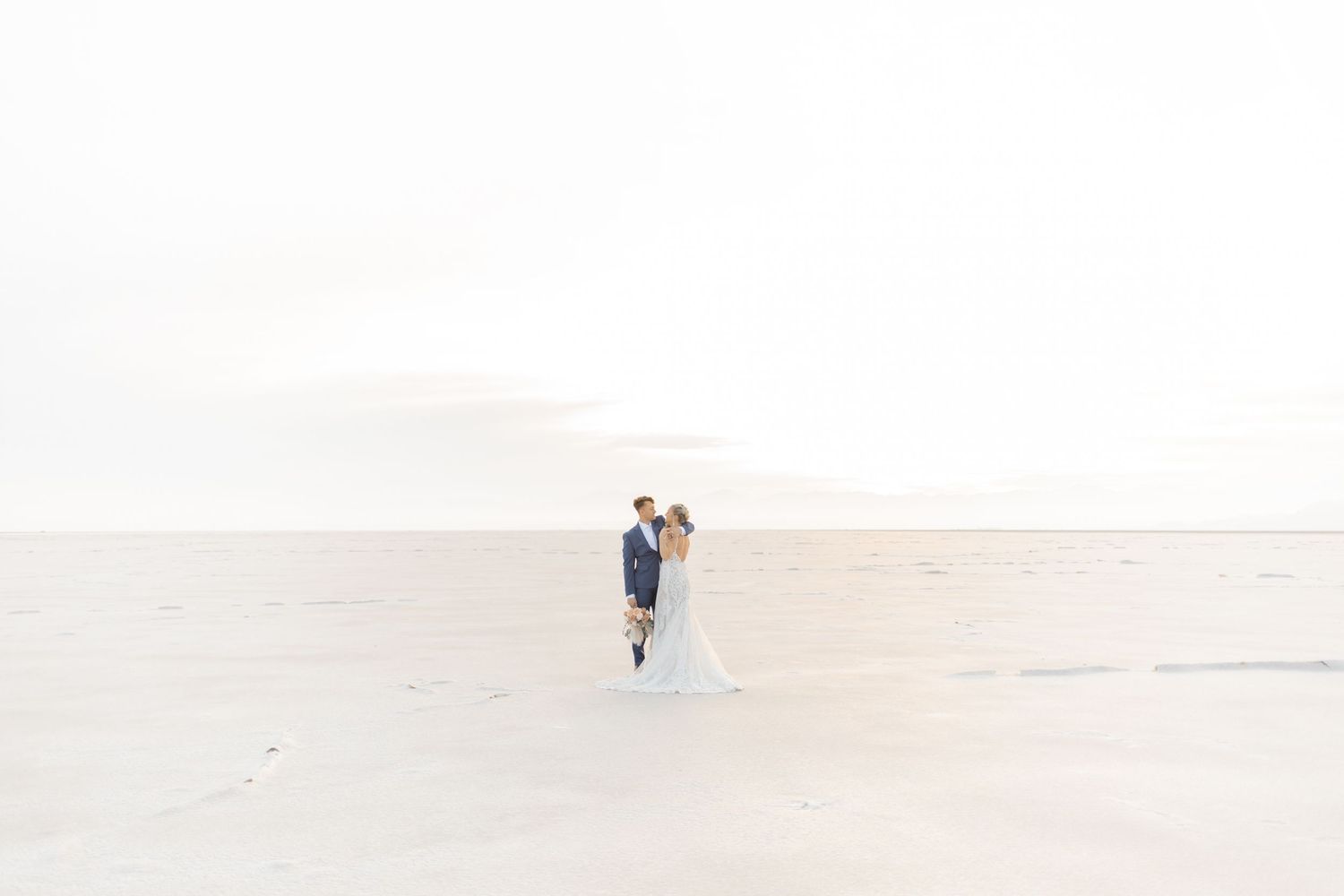 A couple in wedding attire stands together on a white sandy landscape during a desert photoshoot.