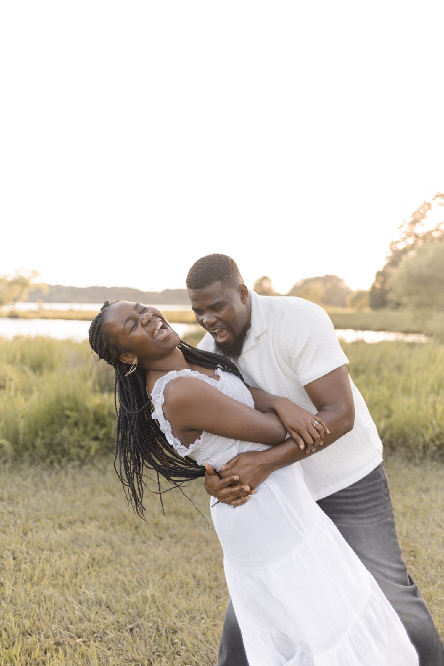 A joyful couple embraces and dances in a sunlit field, the woman in a white dress laughing as the man holds her close.