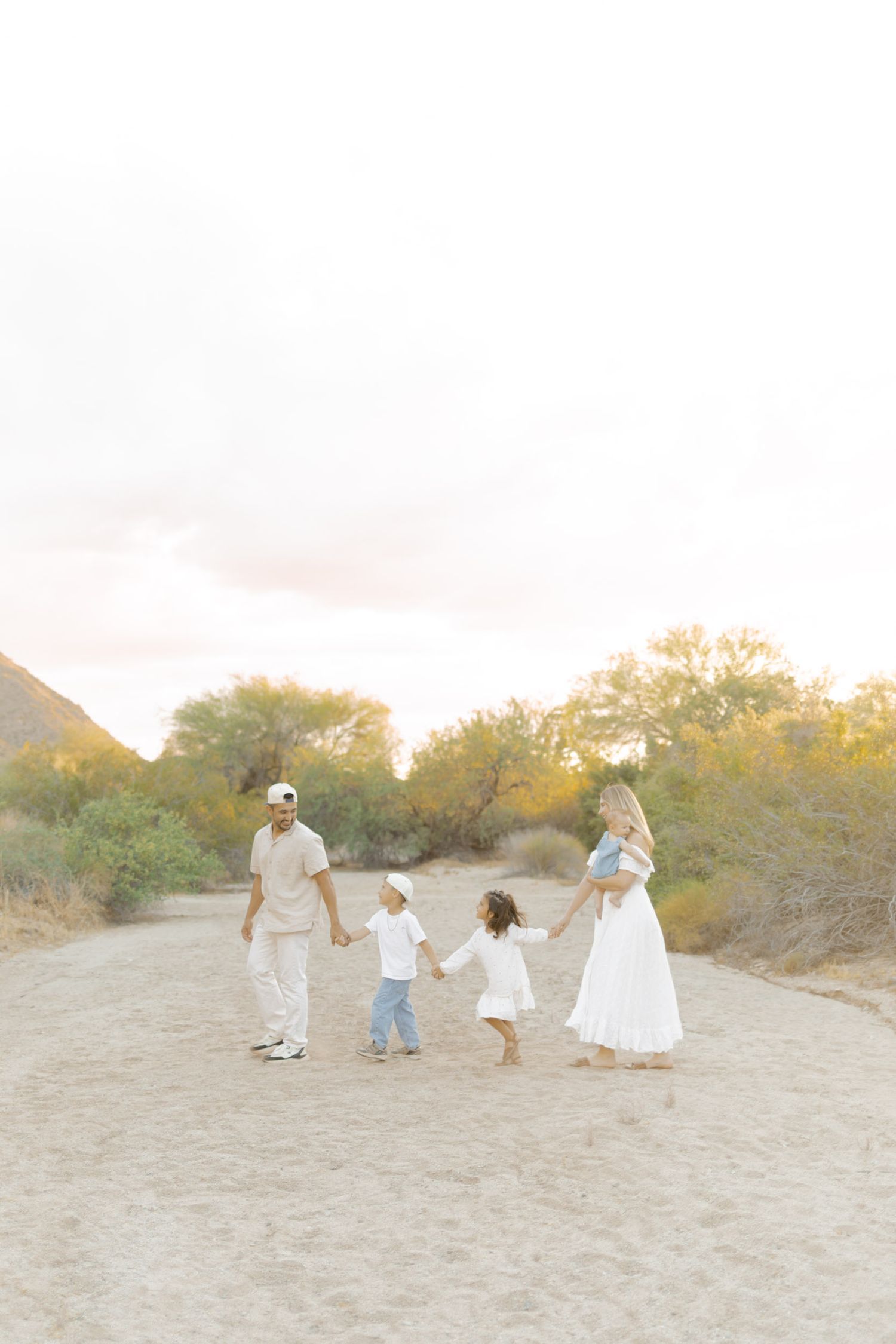 Family of four walking hand in hand on sandy desert path at golden hour in neutral tones.