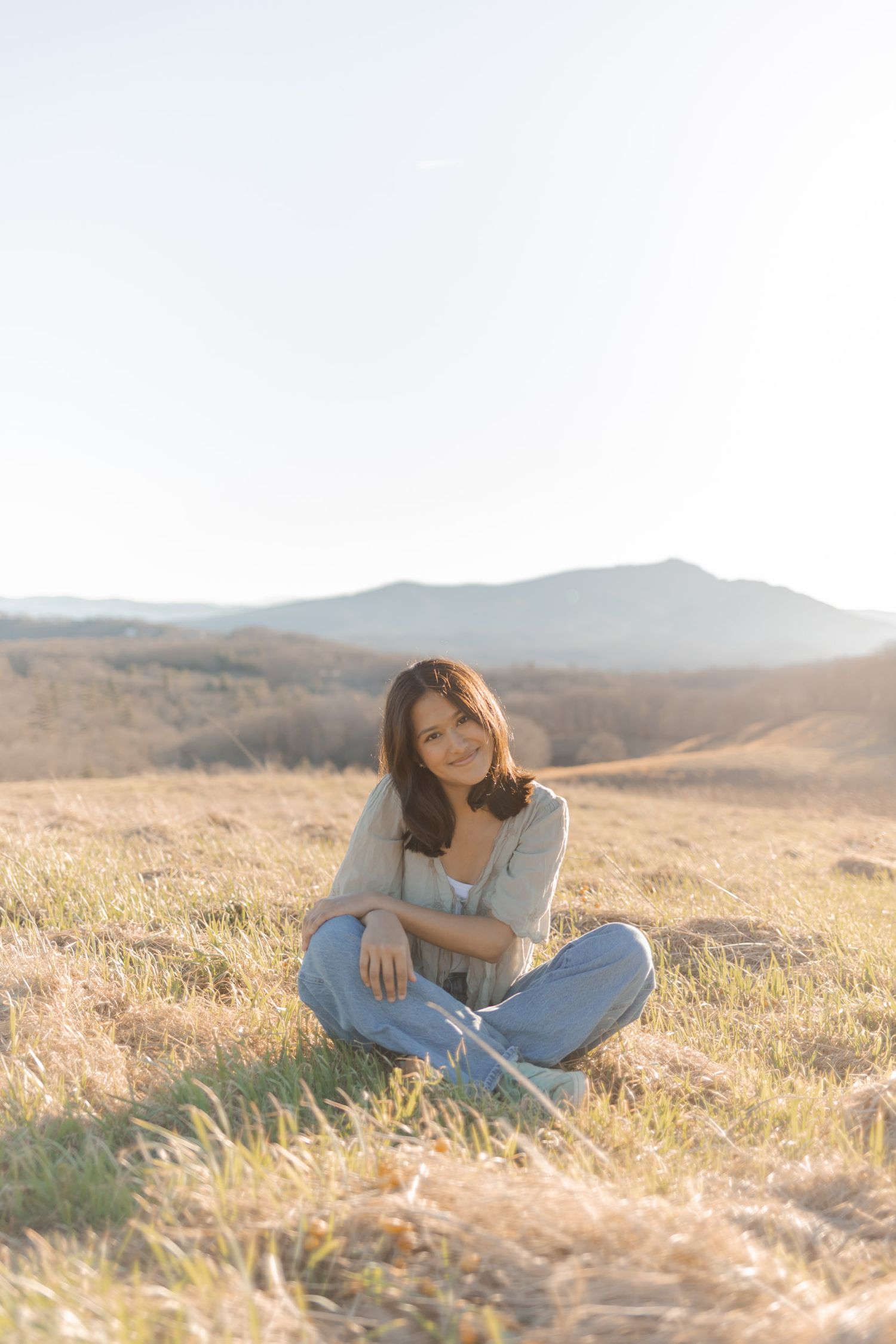 Young woman sitting cross-legged on golden grass field with mountain landscape backdrop in warm sunlight.