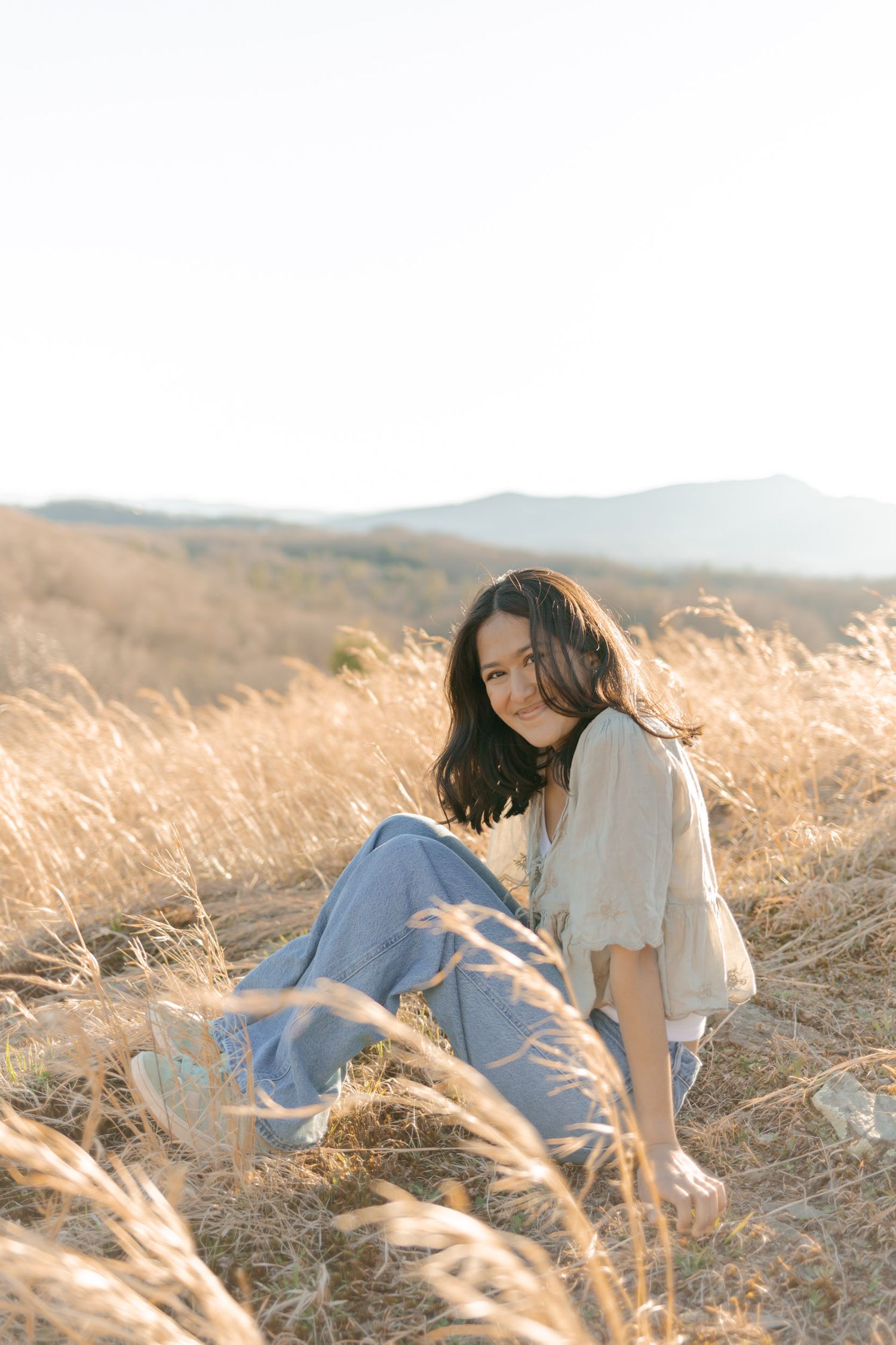 Smiling pregnant woman sitting in golden dry grass field at sunset with mountain landscape in background.