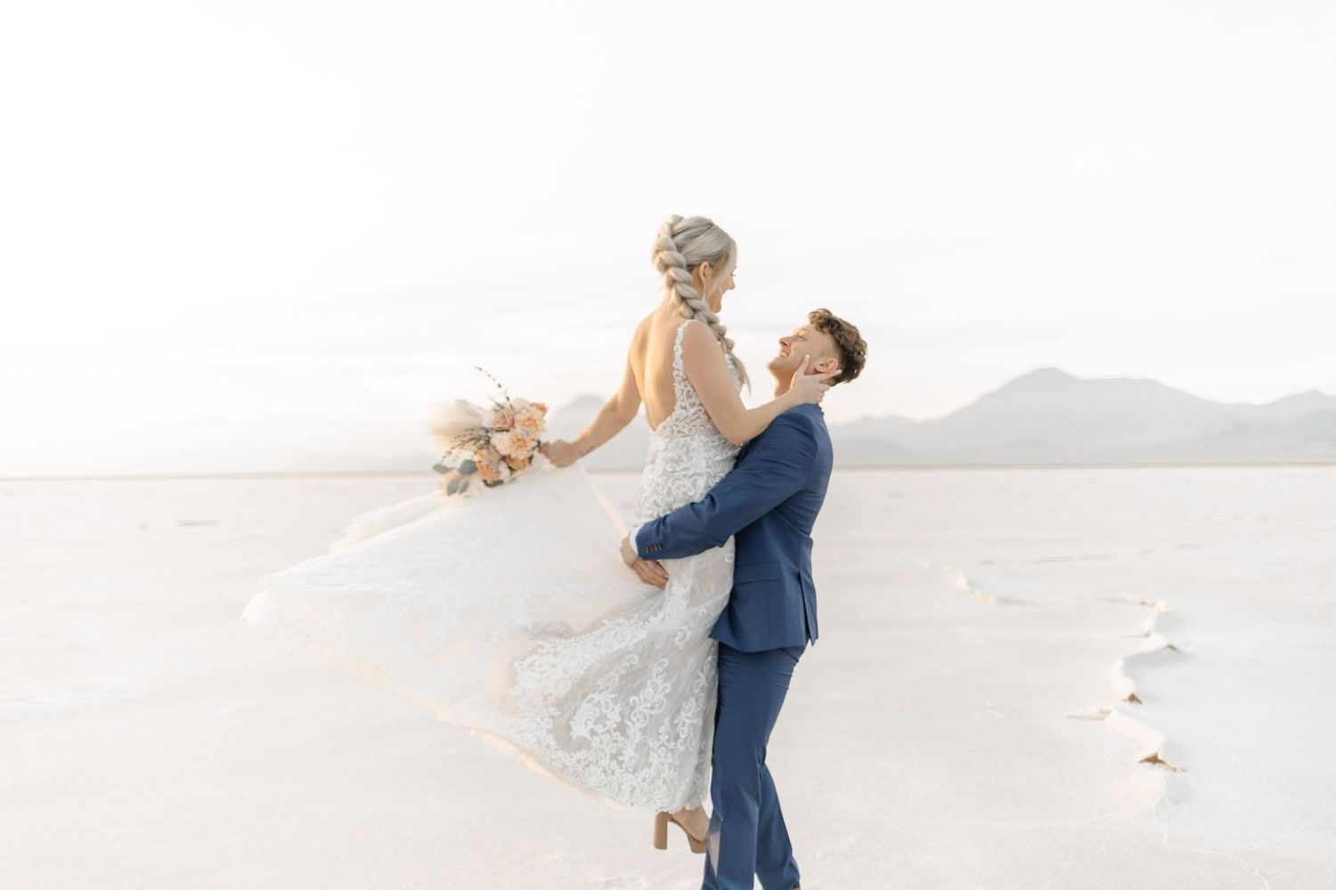 Couple in wedding attire dances and spins together on a scenic white sand beach.