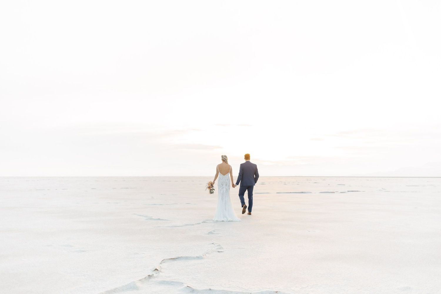 A couple walks hand in hand along a serene white beach shoreline at sunset.
