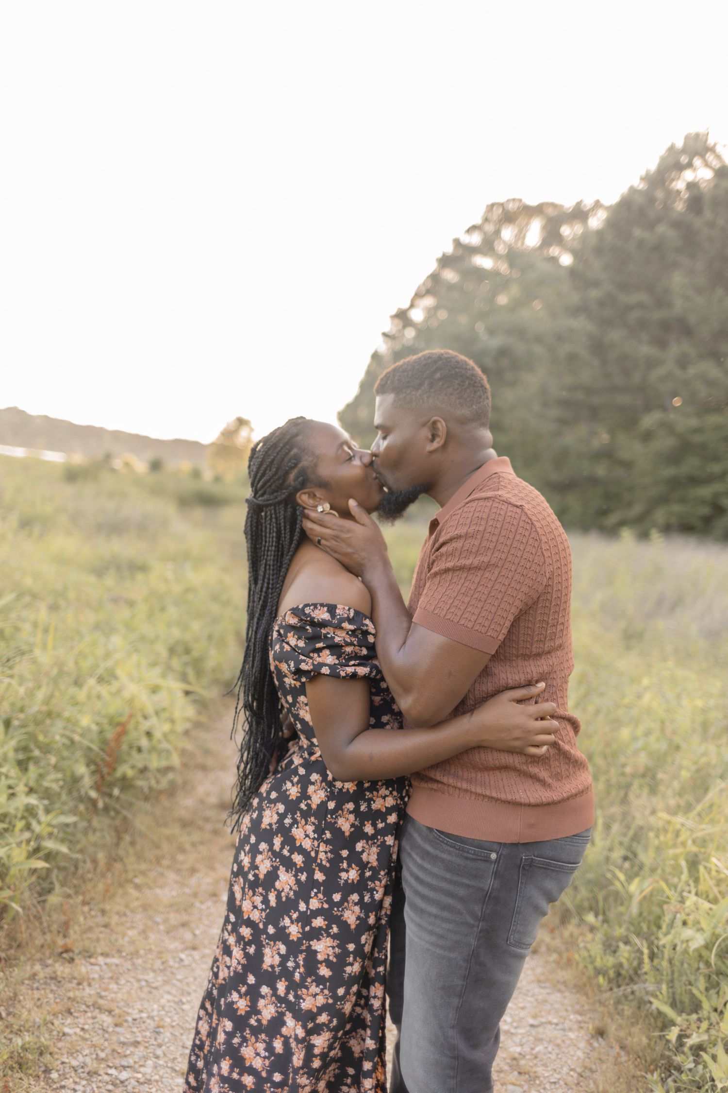 A romantic couple shares a tender kiss on a nature trail surrounded by green fields during golden hour.