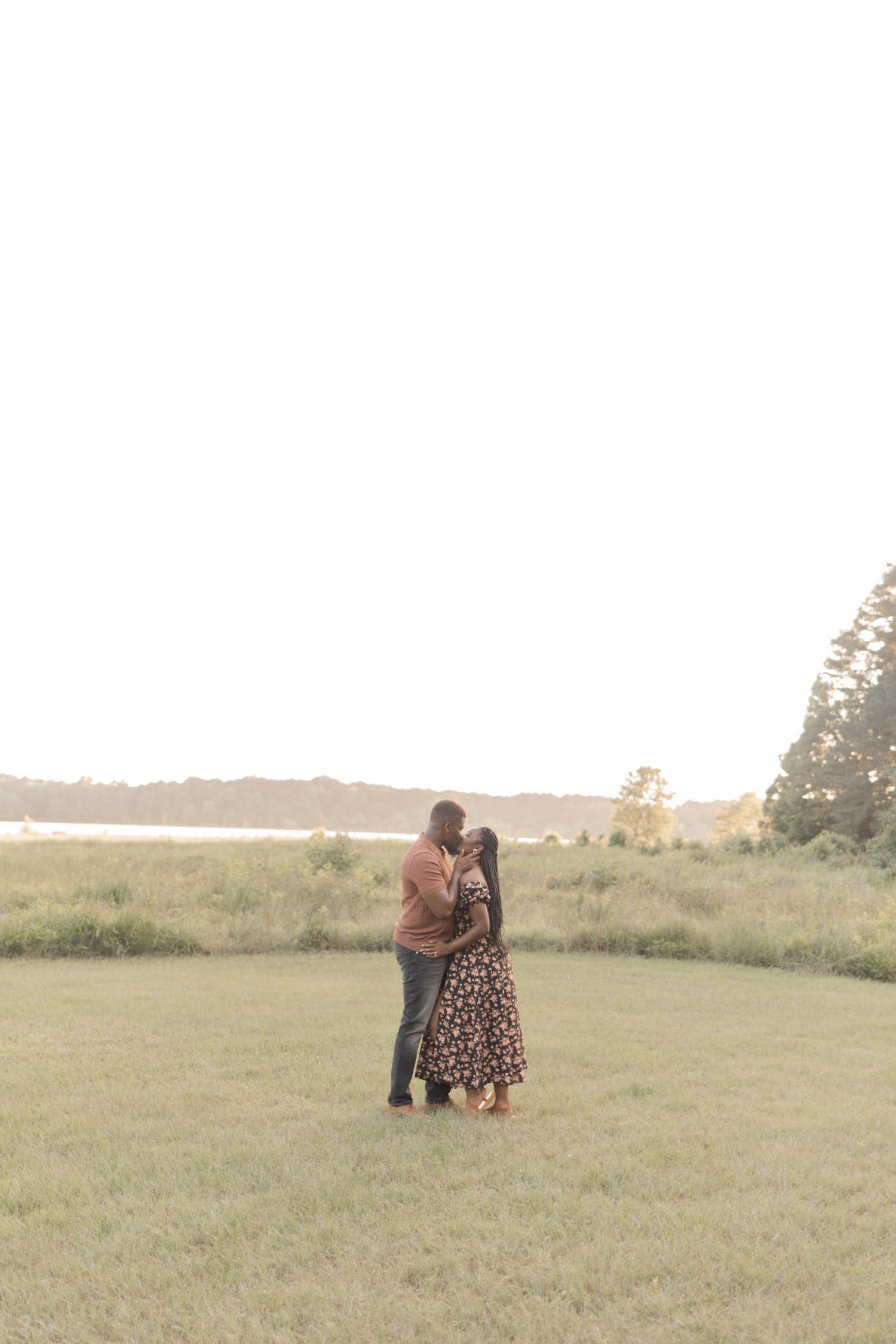 Couple sharing a romantic kiss at golden hour in a lush green field with water and trees in the background.